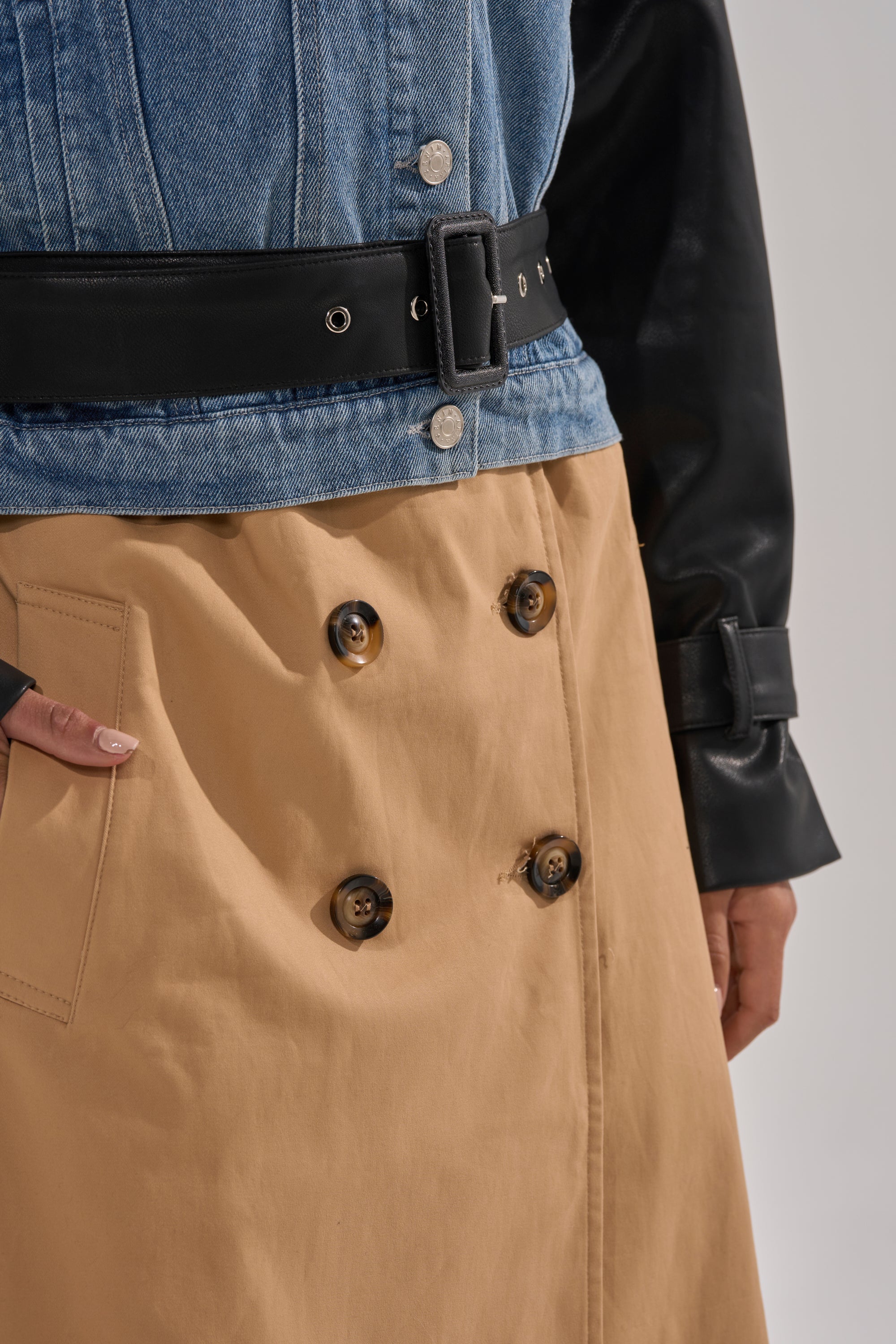 Wearing the CITY SLICKER MIXED MEDIA TRENCH—a denim jacket with faux leather sleeves and a black belt—paired with a tan skirt featuring large buttons and a side pocket, their hand rests casually by the skirt’s pocket.