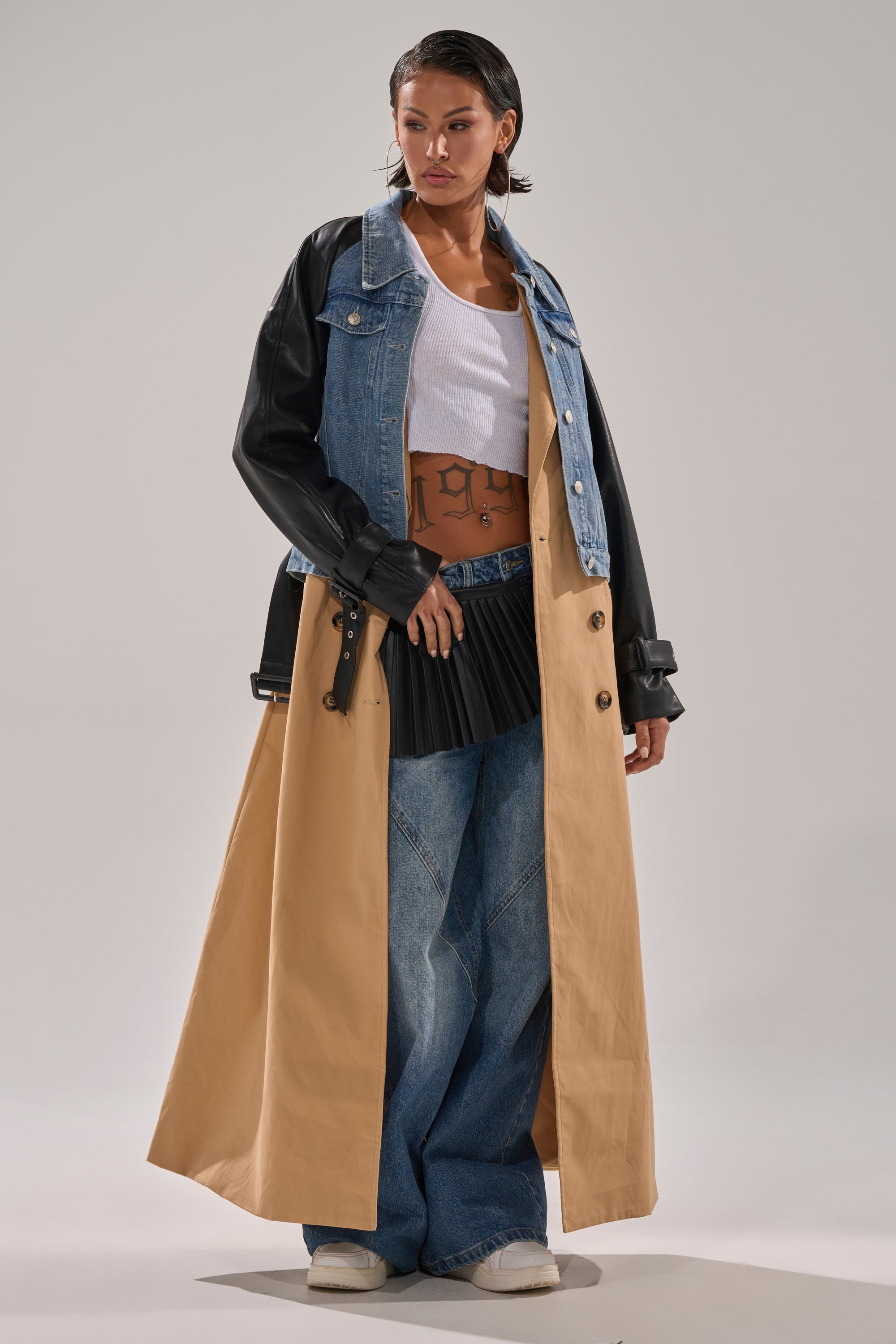 A person poses in a CITY SLICKER MIXED MEDIA TRENCH layered over a white crop top, wide-leg blue jeans, and white sneakers, finished with wet-styled short hair and bold makeup.