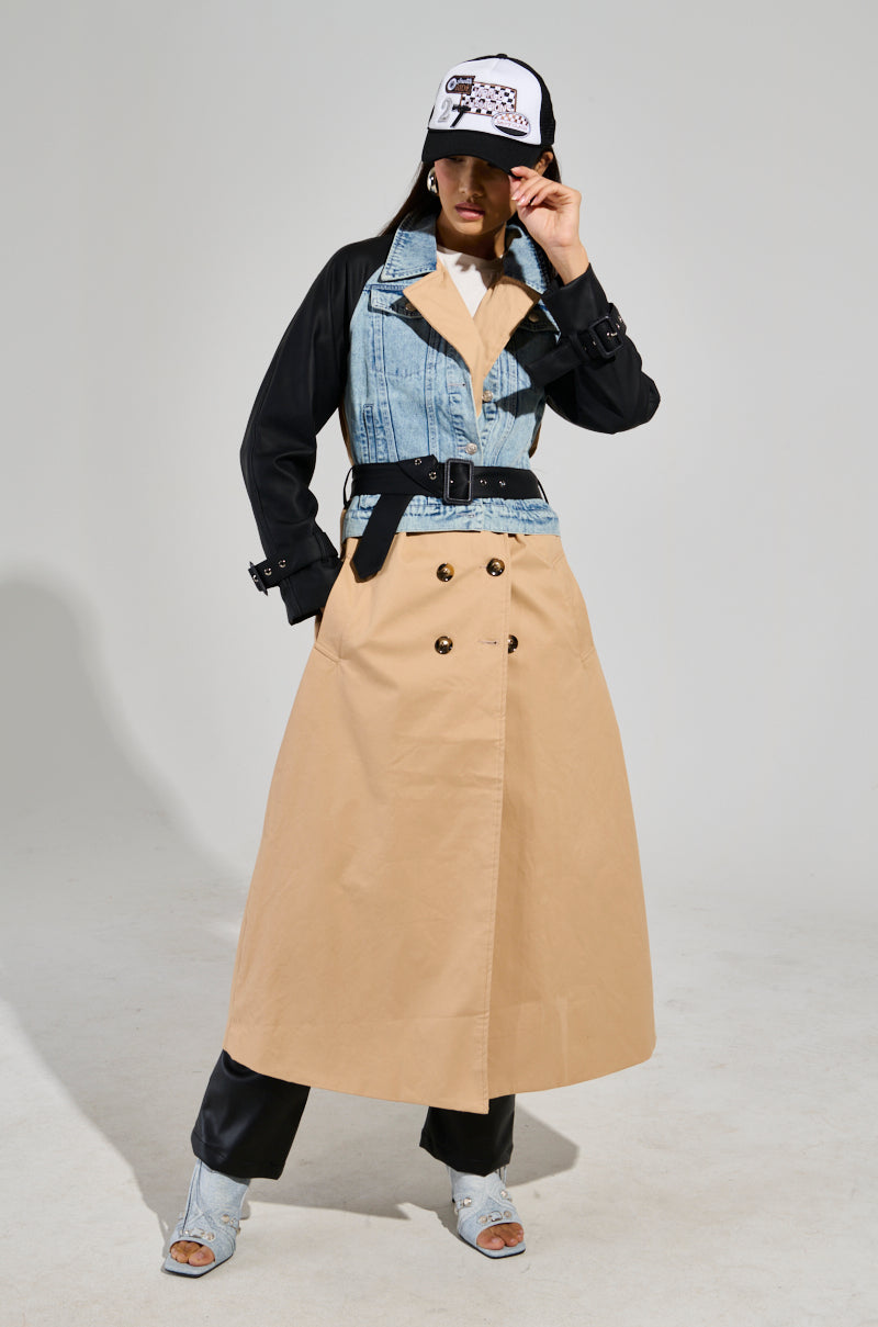 A person is striking a pose in fashionable clothing, featuring the CITY SLICKER MIXED MEDIA TRENCH over black pants and light-colored boots. They wear a logo cap, adding an urban touch against a plain backdrop.