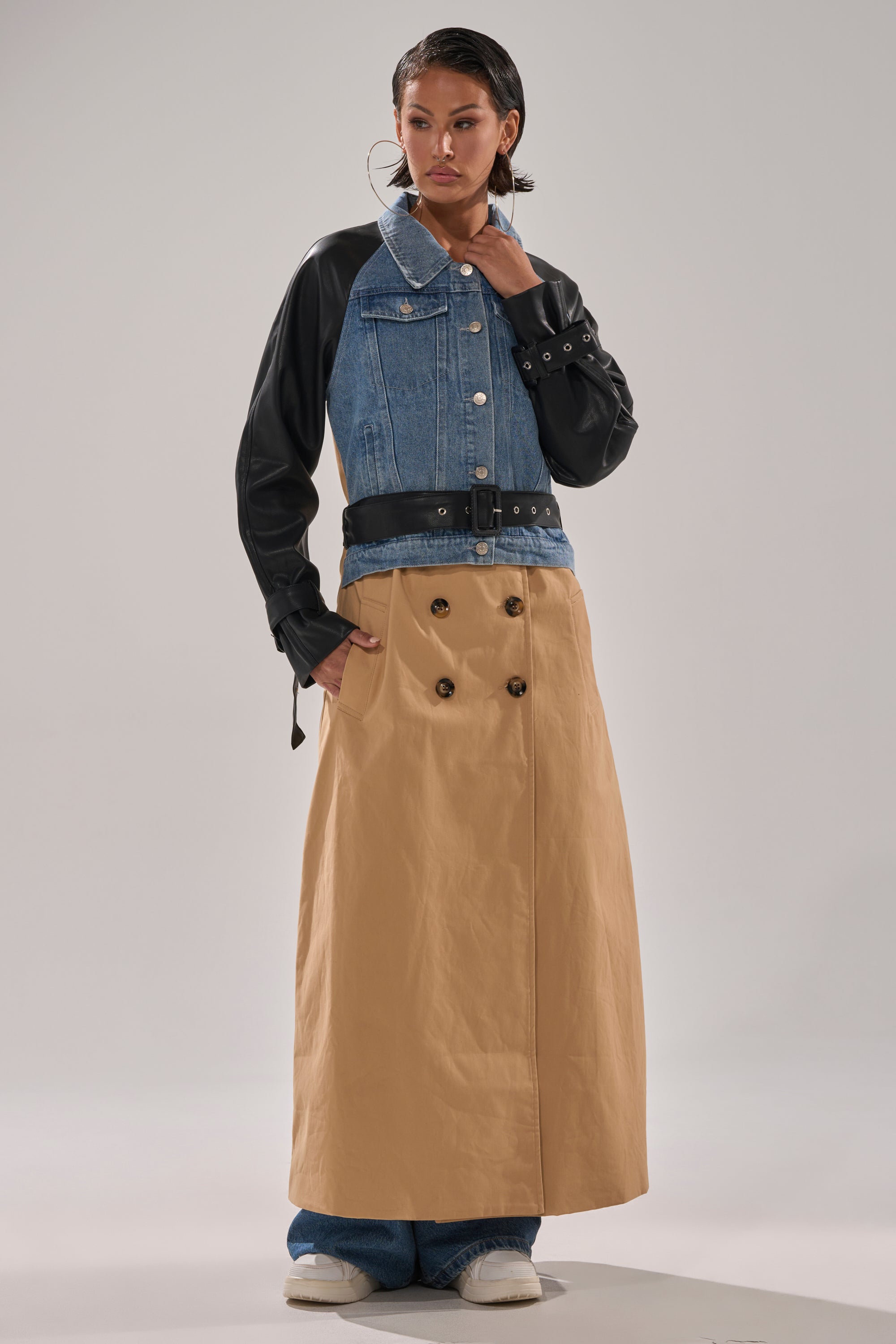 A person poses in a CITY SLICKER MIXED MEDIA TRENCH with black leather sleeves, a wide belt, beige double-breasted skirt detail, blue jeans, white shoes, and large hoop earrings.