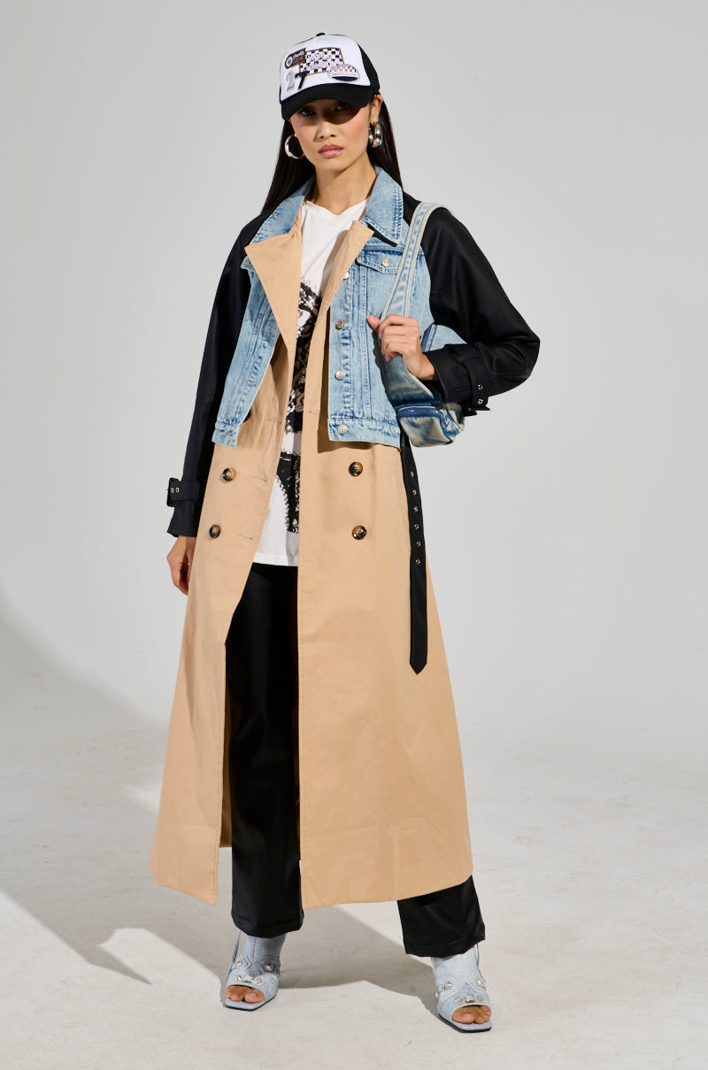 A person stands wearing a City Slicker Mixed Media Trench under a Denim Jacket Overlay, paired with black pants and heeled sandals. They top off the outfit with a white cap and carry a small bag against the plain gray background.