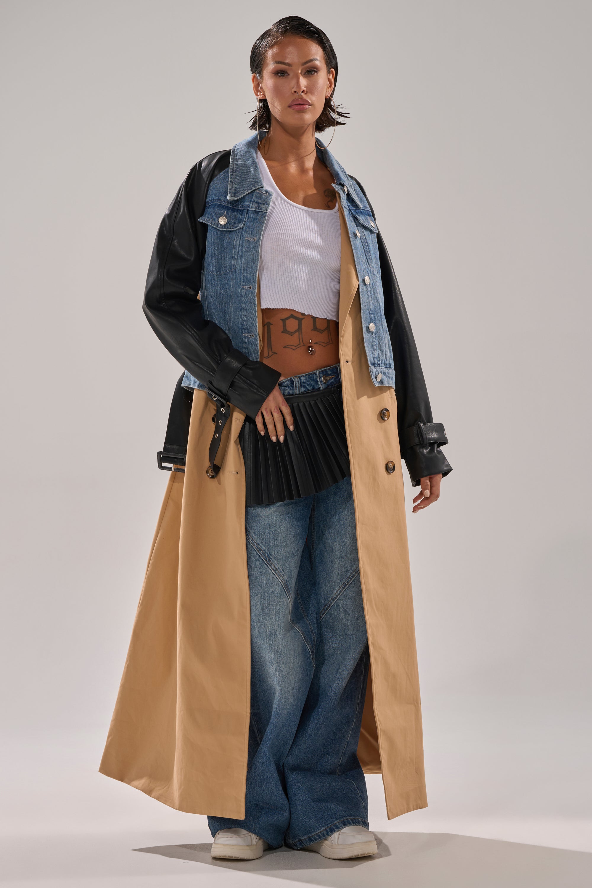 A person models the CITY SLICKER MIXED MEDIA TRENCH with faux leather sleeves and denim overlay, paired with a white tank, wide-leg blue jeans, statement earrings, white shoes, and wet-look hair.