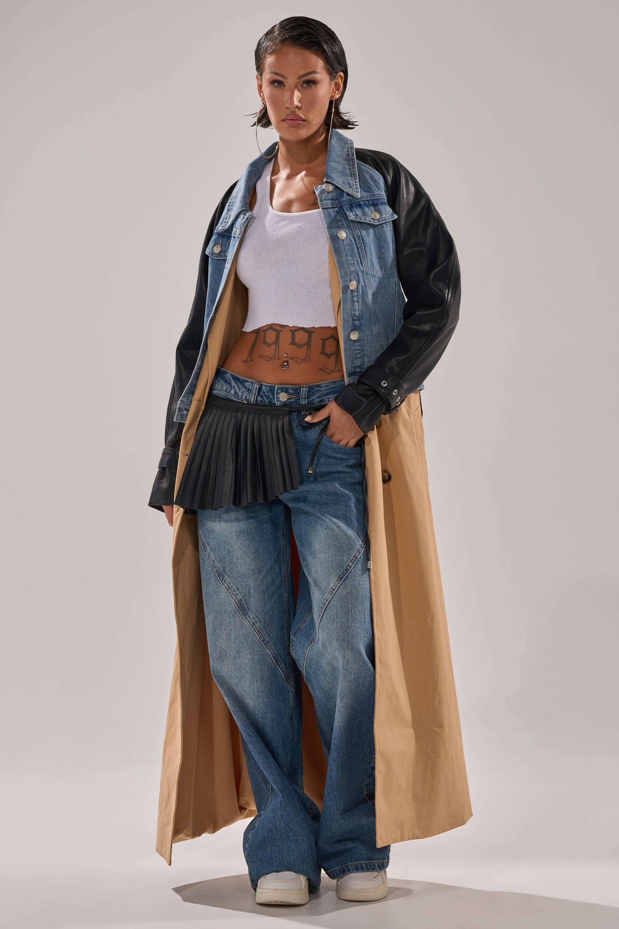 Wearing the CITY SLICKER MIXED MEDIA TRENCH with denim and faux leather sleeves, a white tank top, blue baggy jeans, a black mini skirt over jeans, and white sneakers, a person stands confidently against a plain background.