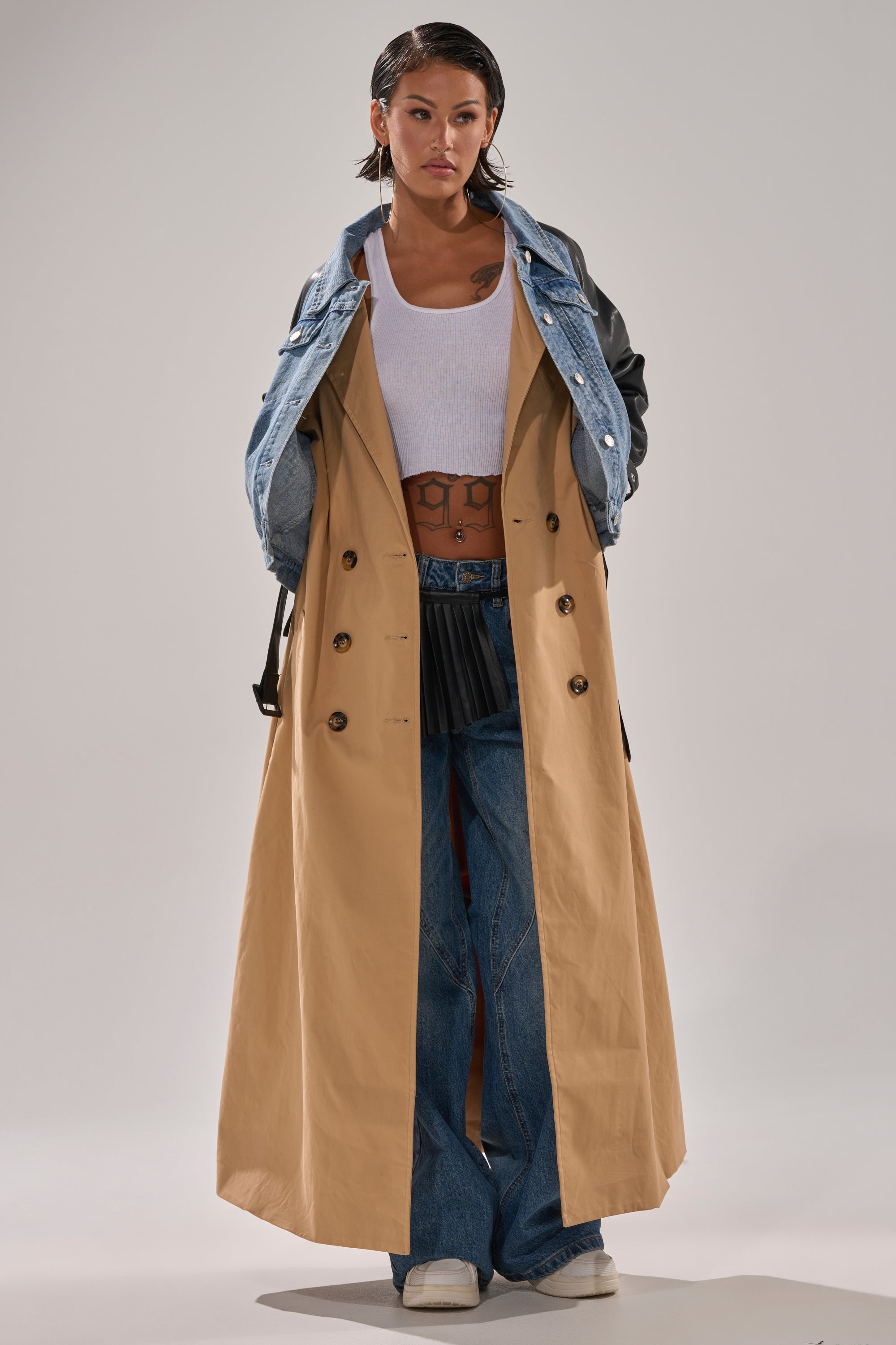 A person stands confidently in the CITY SLICKER MIXED MEDIA TRENCH with faux leather sleeves over a denim jacket overlay, paired with a white tank top, blue jeans, and white shoes. Their slicked-back hair and visible chest and stomach tattoos complete the look.