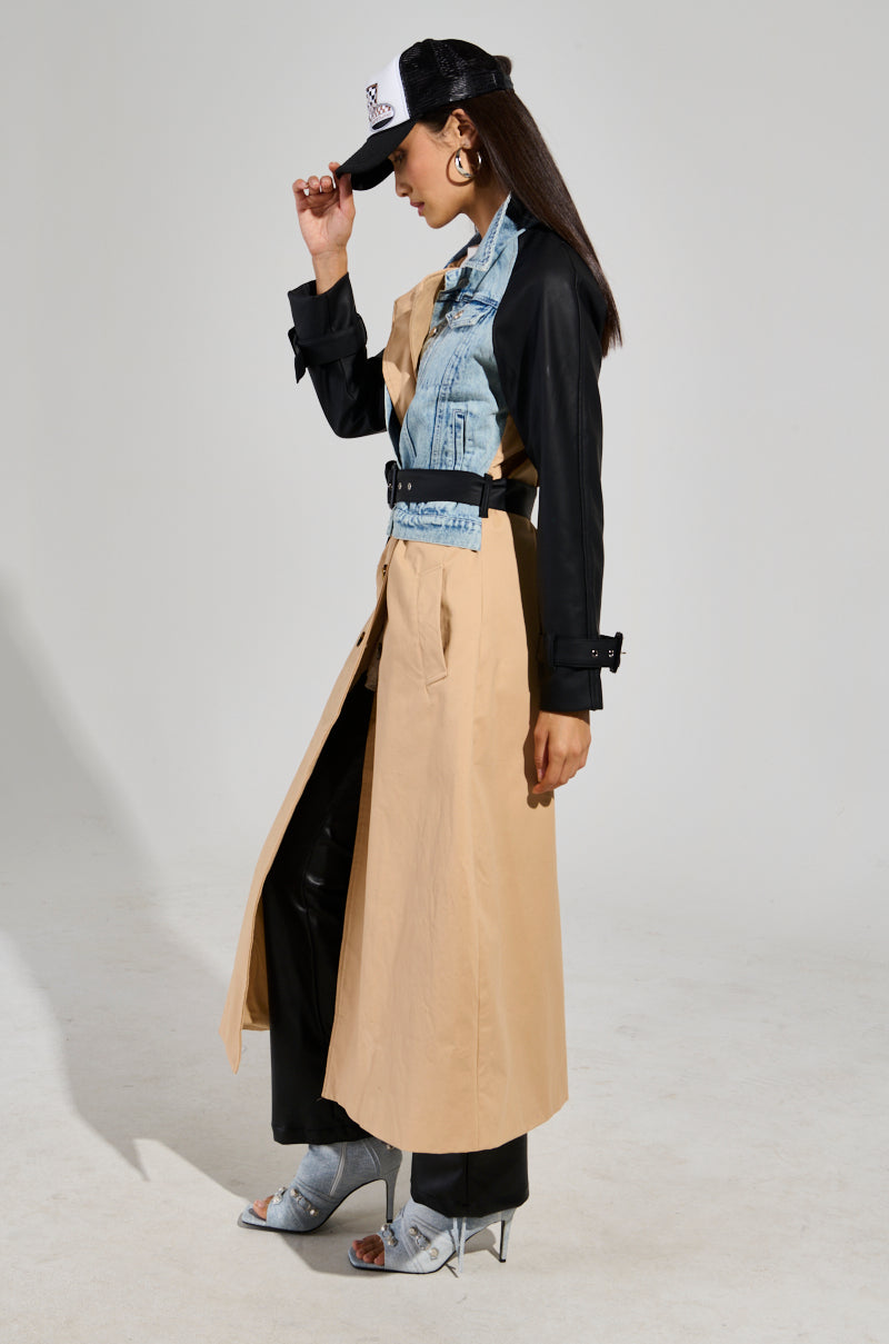 A person wearing a CITY SLICKER MIXED MEDIA TRENCH over a black shirt and trousers, complemented by silver high-heeled shoes. They're adjusting a black and white cap, standing in side profile against a plain background.