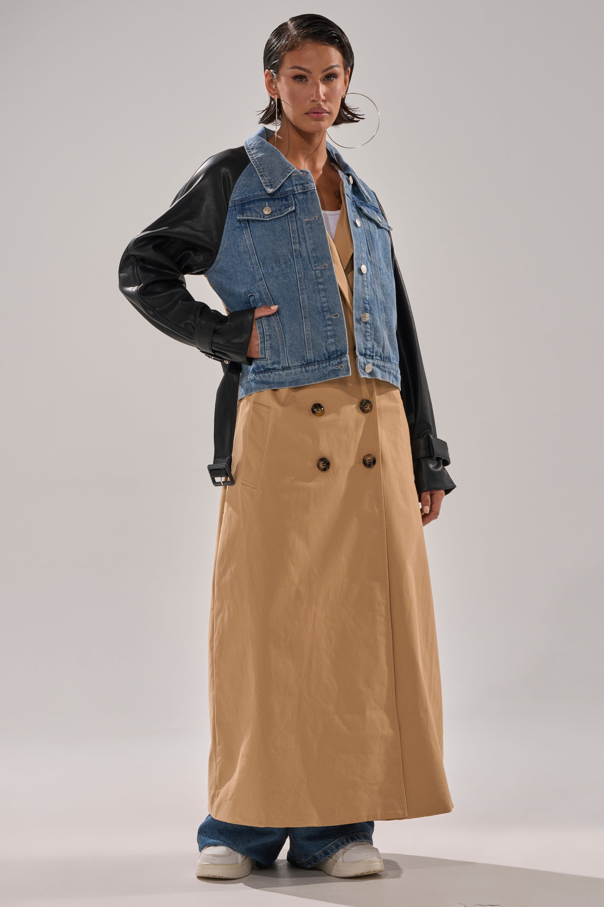 A person wears the CITY SLICKER MIXED MEDIA TRENCH over blue jeans, styled with a cropped denim jacket with faux leather sleeves, wet-look hair, hoop earrings, and white shoes.