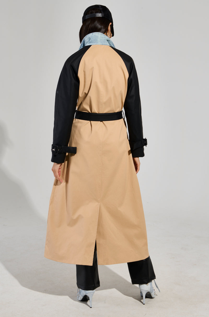 A person is wearing a CITY SLICKER MIXED MEDIA TRENCH, featuring a denim jacket overlay with black sleeves. They complement it with black pants and light-colored shoes. Facing away and adorned in a black hat, they stand against a plain white background.