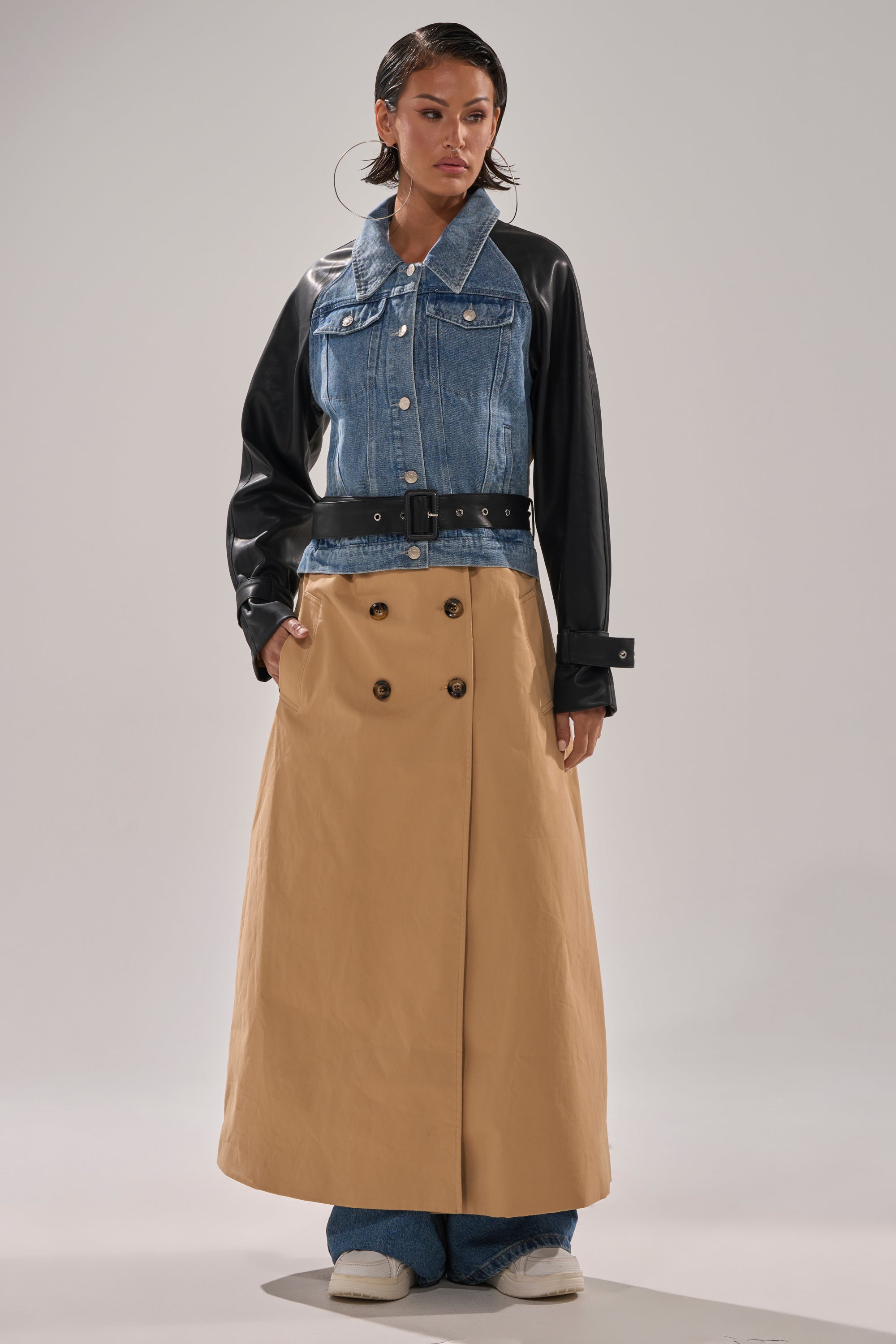 A woman models the CITY SLICKER MIXED MEDIA TRENCH—a blue denim jacket with faux leather sleeves—styled with a wide black belt, tan double-breasted maxi skirt, blue jeans, and white shoes against a plain background.