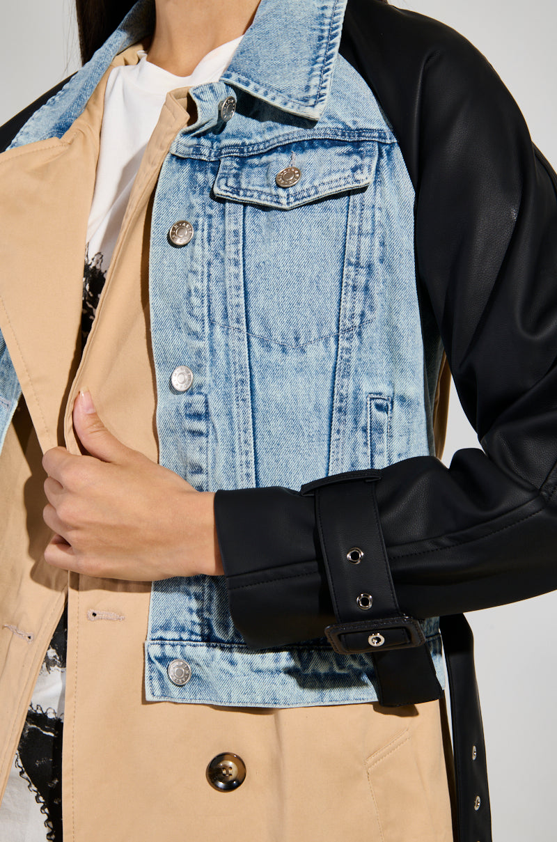 The individual is dressed in the CITY SLICKER MIXED MEDIA TRENCH, which showcases a light blue denim panel combined with beige fabric and black leather sleeves. The coat is slightly open at the front, offering a peek of a white shirt underneath, while they adjust the jacket with one hand.