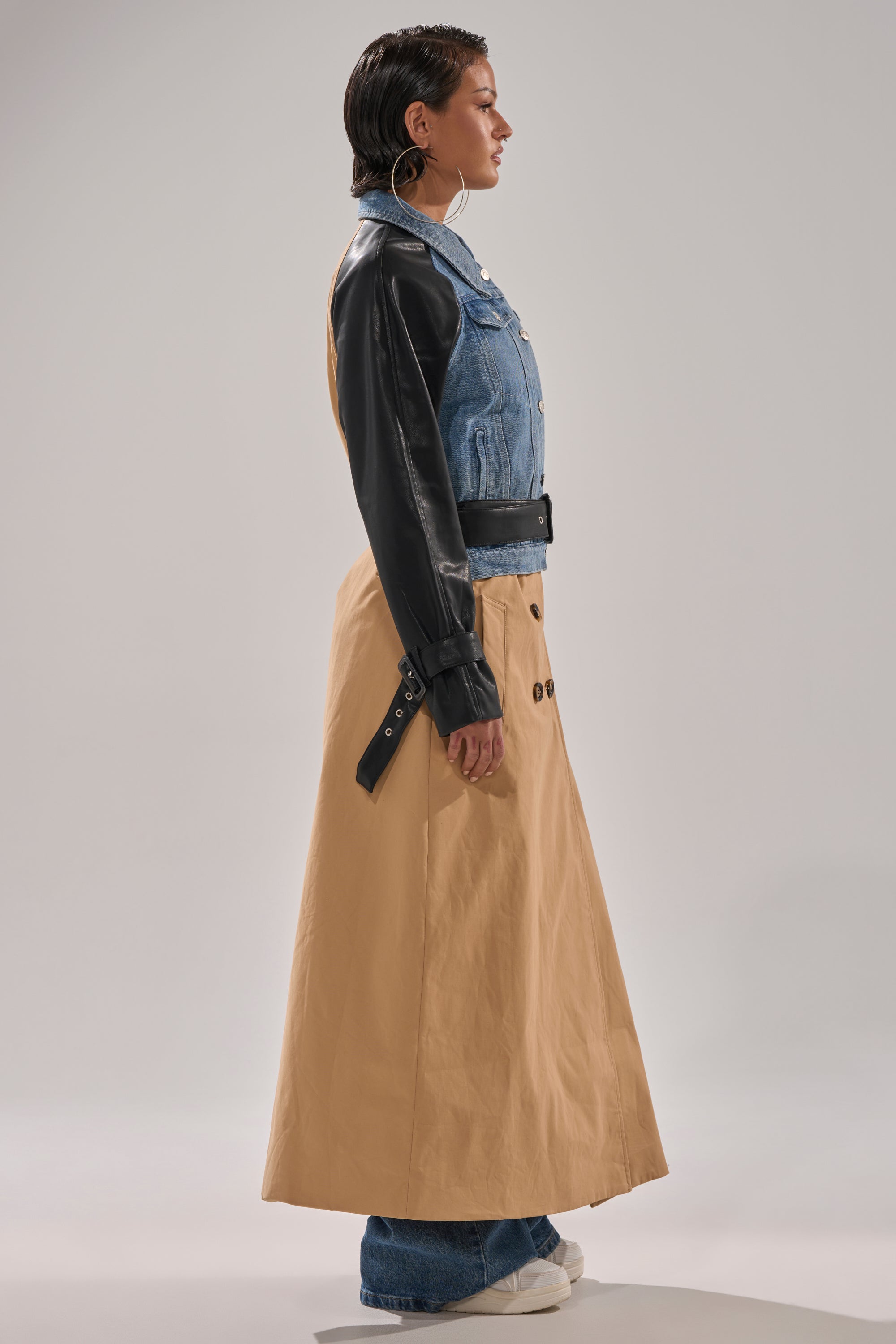 A woman in profile wears the CITY SLICKER MIXED MEDIA TRENCH, styled with a long tan skirt over blue jeans and white sneakers, set against a plain light background.