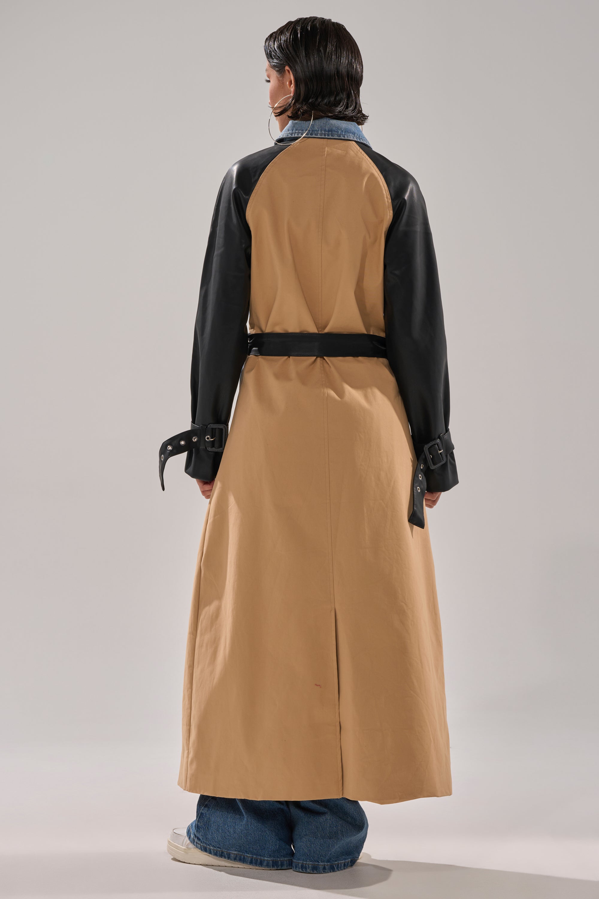 A person stands facing away in the CITY SLICKER MIXED MEDIA TRENCH, featuring tan fabric with faux leather sleeves and a black belt, paired with blue jeans and white sneakers against a plain light background.