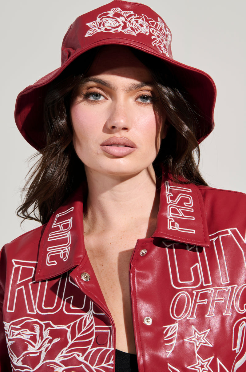 A person with long hair sports the ROSE CITY EMBROIDERED PU BUCKET HAT in red and a matching jacket, both adorned with white graphic designs and text, as they face forward against a neutral background. 0