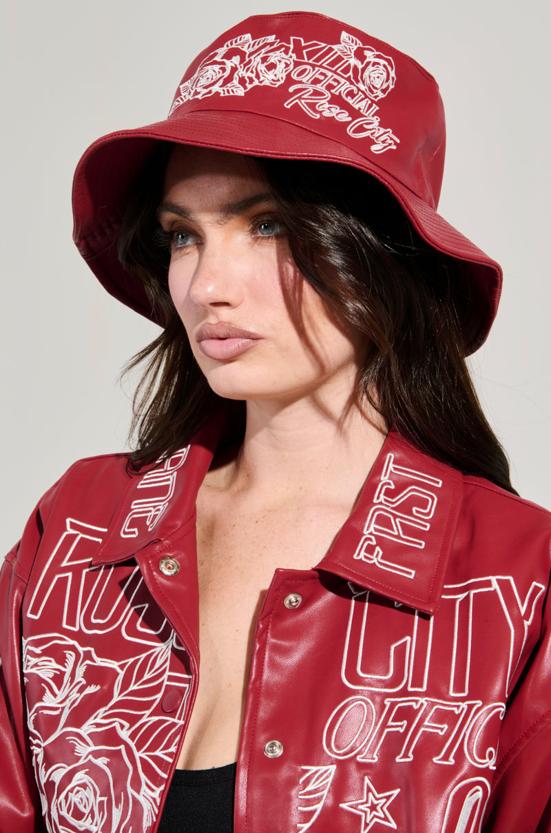 A woman wears a red ROSE CITY EMBROIDERED PU BUCKET HAT and a matching jacket adorned with white floral motifs and words like "OFFICIAL" and "CITY." She has long dark hair, a neutral expression, standing against a plain background. 1