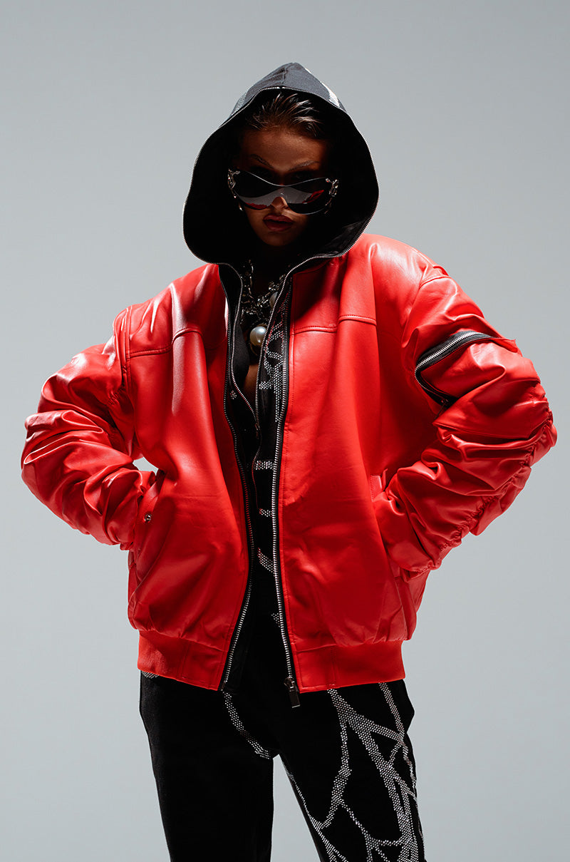 Person wearing the STORMY OVERSIZED BOMBER in a shiny red finish, with the hood pulled up, complemented by dark sunglasses and black pants featuring white designs. Their confident pose with hands on hips against a plain background accentuates the bold style of the oversized silhouette.