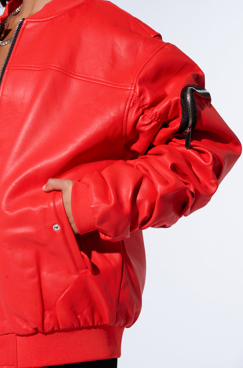A person is shown from the torso up, wearing the STORMY OVERSIZED BOMBER, a vibrant red jacket featuring a zippered pocket on the sleeve and ribbed cuffs. The faux leather jacket boasts a shiny finish and an oversized silhouette. The background is plain and light-colored.