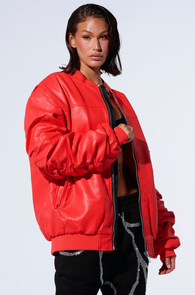 A person with short dark hair wears the STORMY OVERSIZED BOMBER, a large, bright red jacket with an oversized silhouette, over a partially unzipped black top. Stylishly posing against a plain white background, they also sport black pants with white and silver detailing.