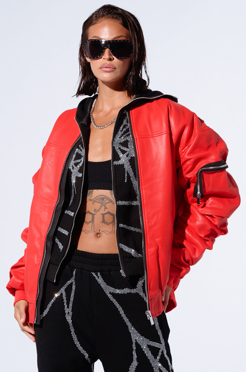 A person with long dark hair wearing large black sunglasses and the STORMY OVERSIZED BOMBER in red faux leather with a black inner lining. They have on a black crop top revealing a tattoo on their upper abdomen and are also wearing black pants with a white abstract design.