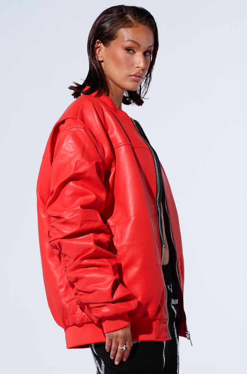 A person with shoulder-length dark hair, wearing the STORMY OVERSIZED BOMBER in bright red faux leather, poses with a serious expression against a plain background. The individual is also dressed in black pants featuring white zippers and is looking slightly over their shoulder.