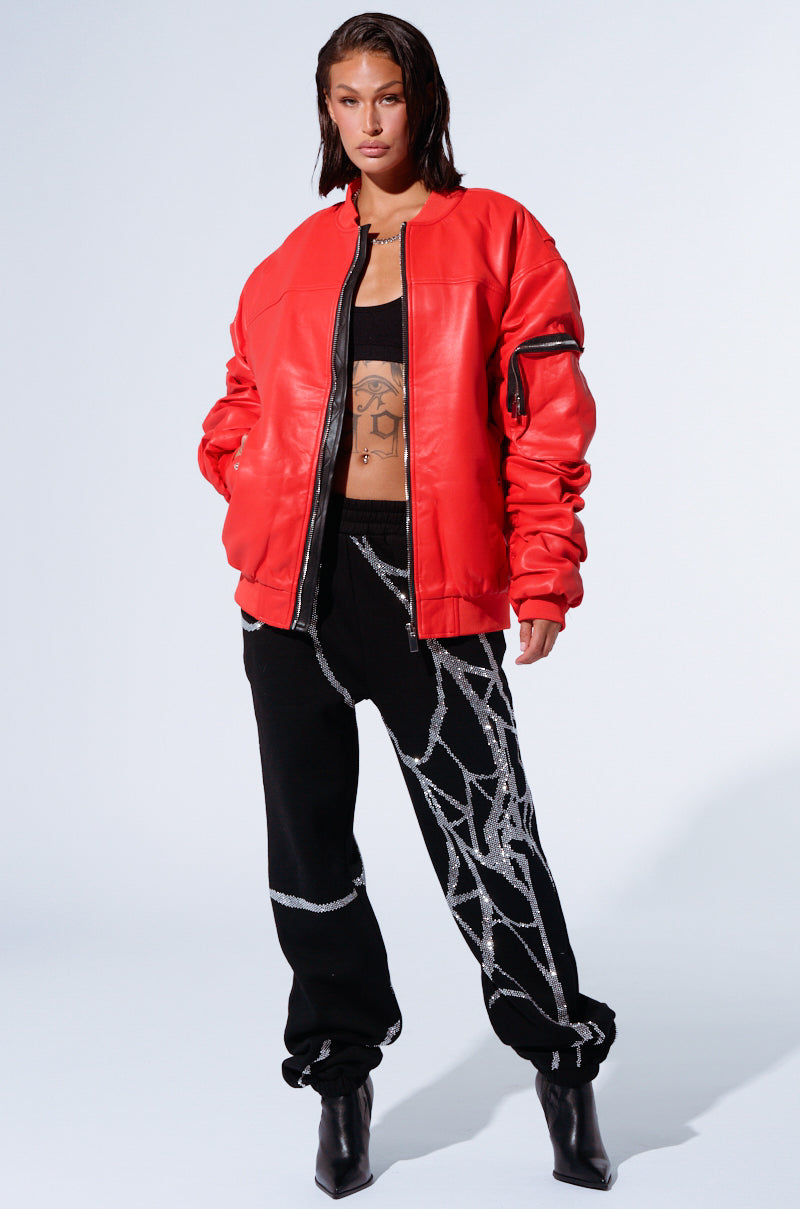 A person is dressed in a stylish combination of the STORMY OVERSIZED BOMBER, a bright red, oversized bomber jacket, and black pants adorned with silver sequin designs. They wear a black crop top underneath the jacket and black heeled boots, standing confidently against a plain background.