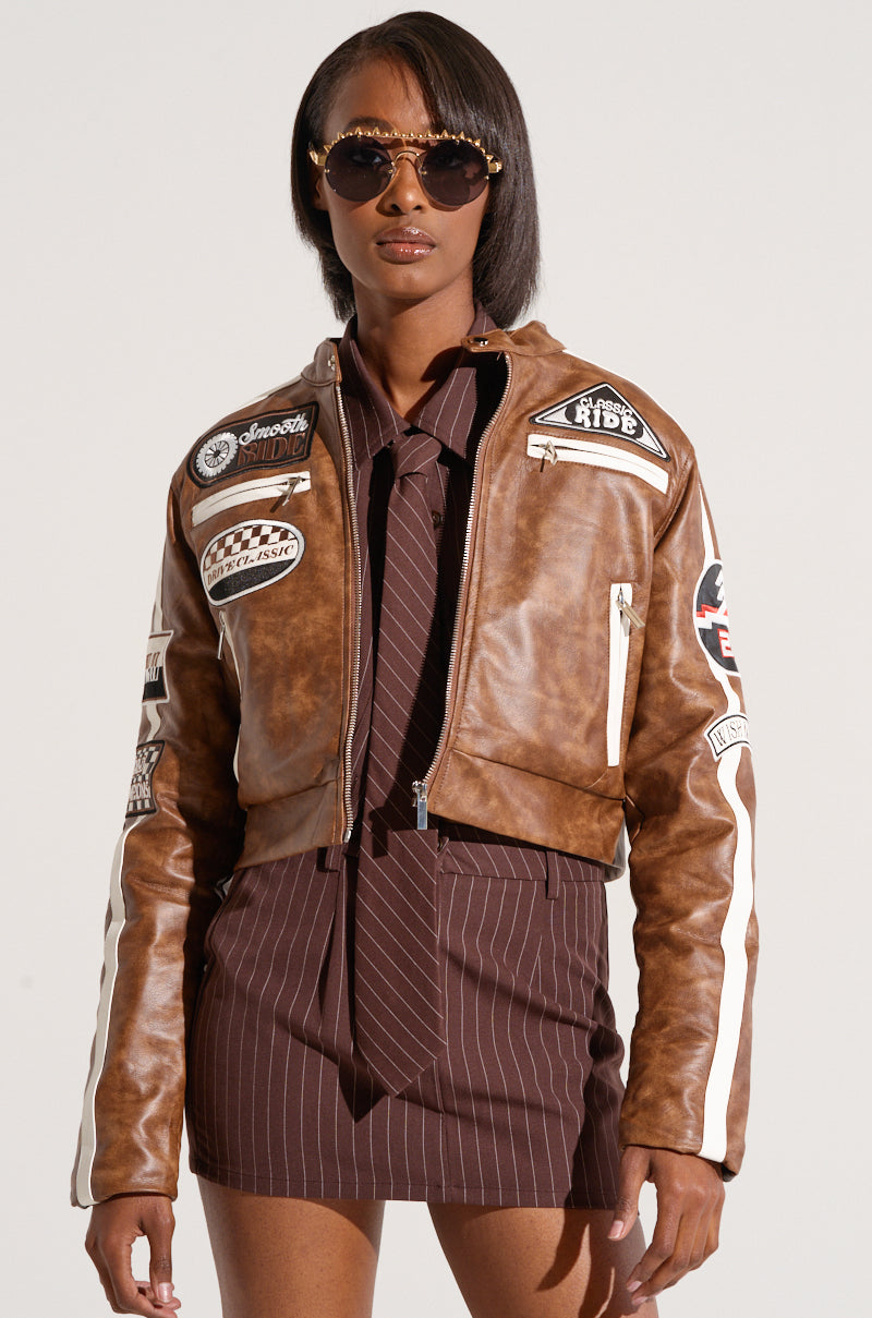 A person wearing sunglasses, a WISH ME LUCK EMBROIDERED MOTO jacket, and a striped brown shirt with a matching tie stands in front of a plain background.