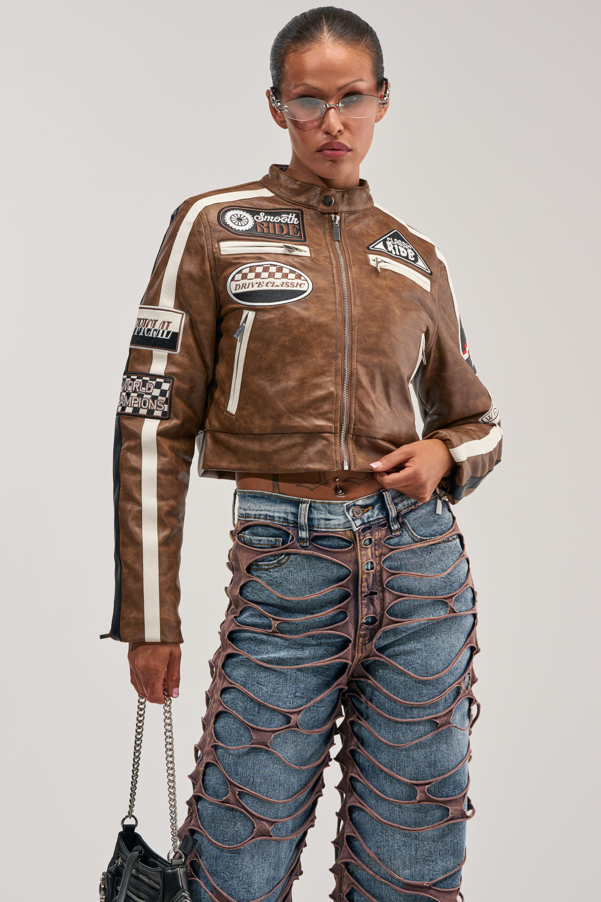 Wearing the WISH ME LUCK EMBROIDERED MOTO—a brown faux leather jacket adorned with racing-inspired patches—this individual pairs it with unique blue jeans featuring large cutouts, accessorizes with a black bag, and poses confidently with one hand on their hip, meeting the camera's gaze.
