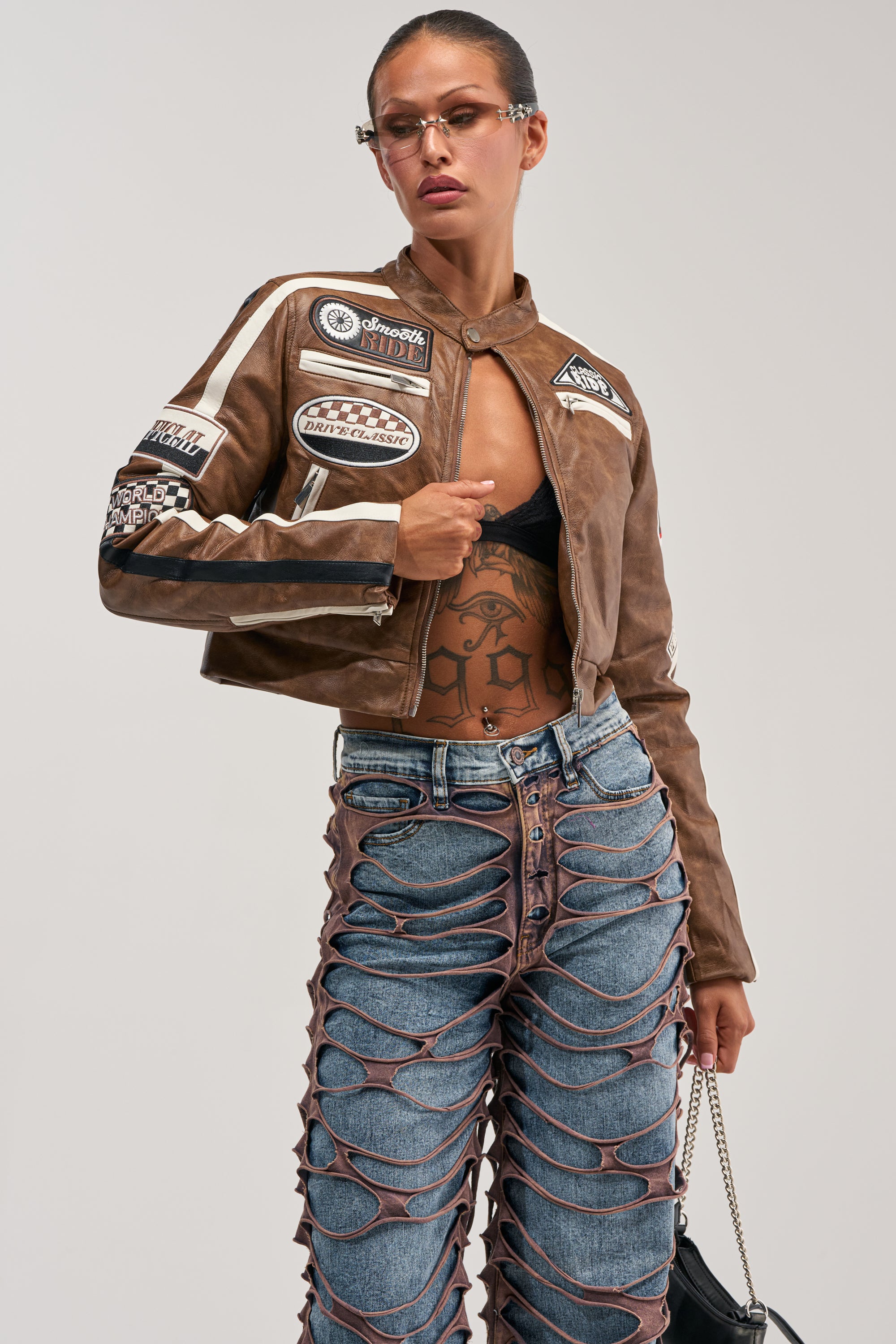 A woman wearing the WISH ME LUCK EMBROIDERED MOTO, paired with ripped blue jeans featuring criss-cross detailing, glasses, and holding a purse poses confidently against a plain background.