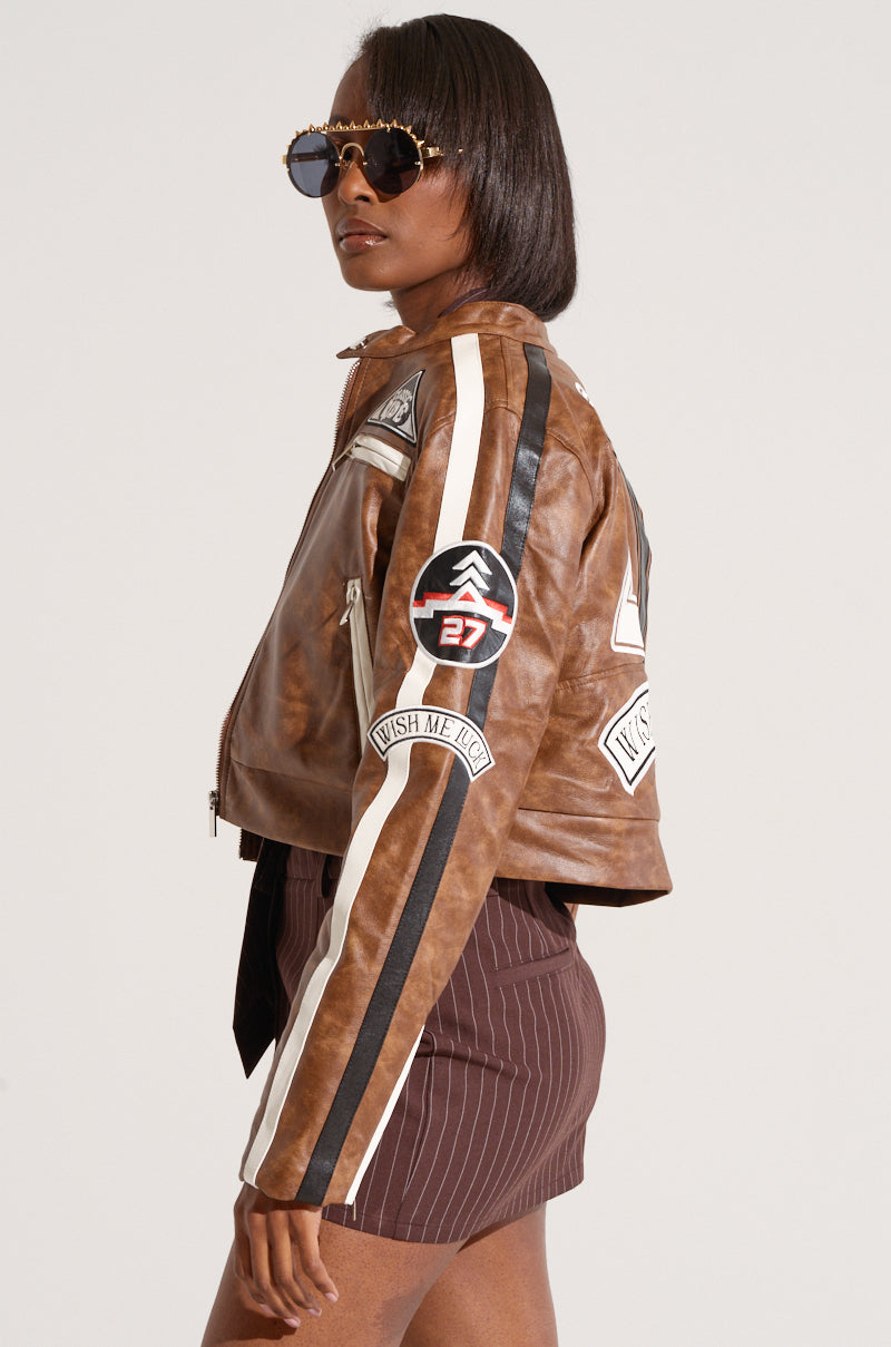 A person models the AKIRA Label WISH ME LUCK EMBROIDERED MOTO brown faux leather jacket, adorned with several patches. They wear dark sunglasses, and the jacket features stripes down the sleeves and a prominent "27" embroidered letterman patch on the arm. The person also wears a short, pinstriped brown skirt.