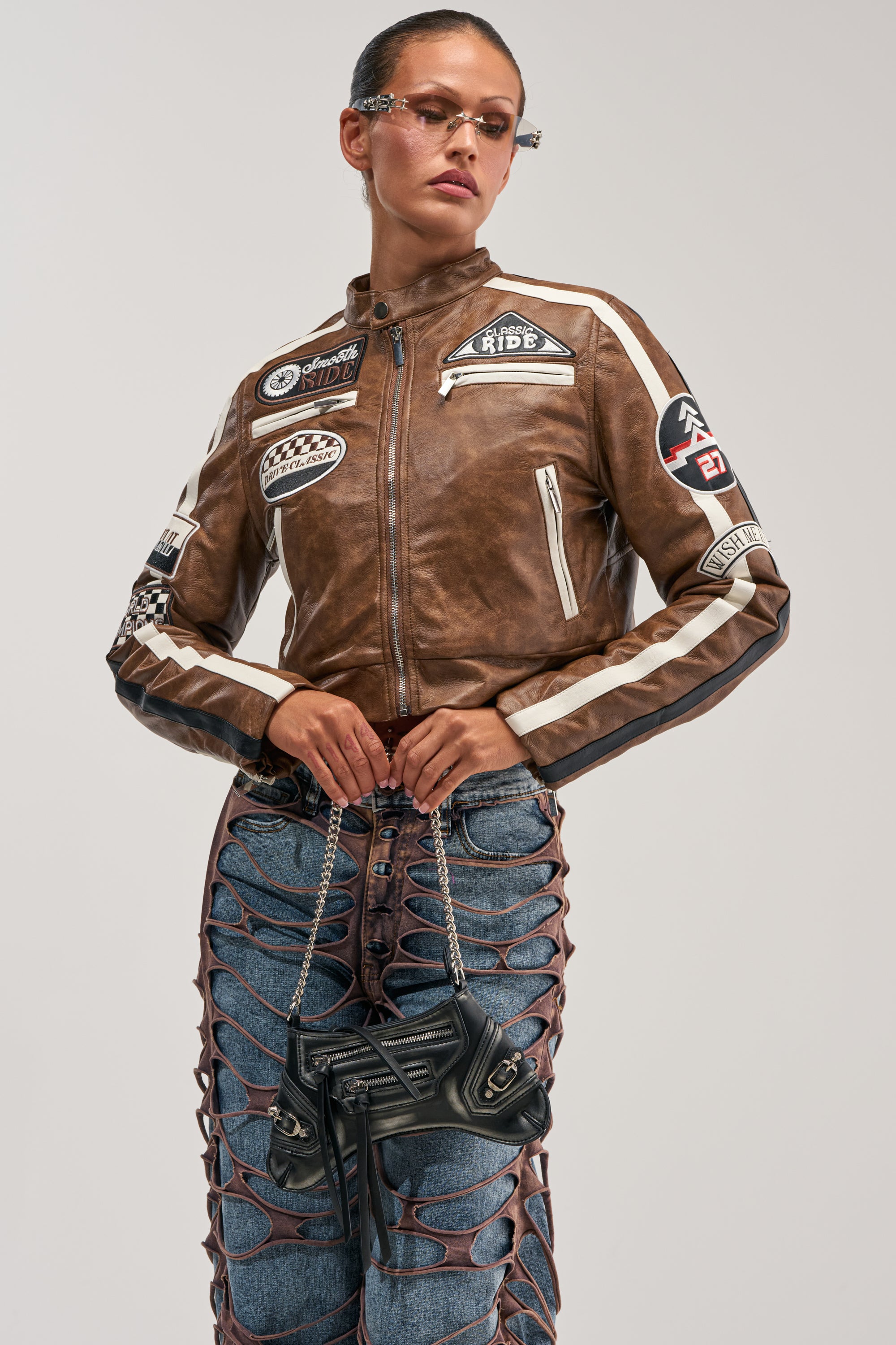 Wearing the WISH ME LUCK EMBROIDERED MOTO jacket, this person styles it with unique jeans featuring large cutouts, a small black bag, slicked-back hair, and narrow sunglasses.
