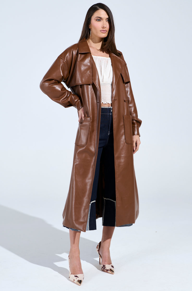 A woman exudes confidence in the Butter Luxe Faux Leather Trench in brown over a white top and dark jeans, paired with patterned pointed shoes. She stands boldly against a plain background, showcasing the trench's elegance.