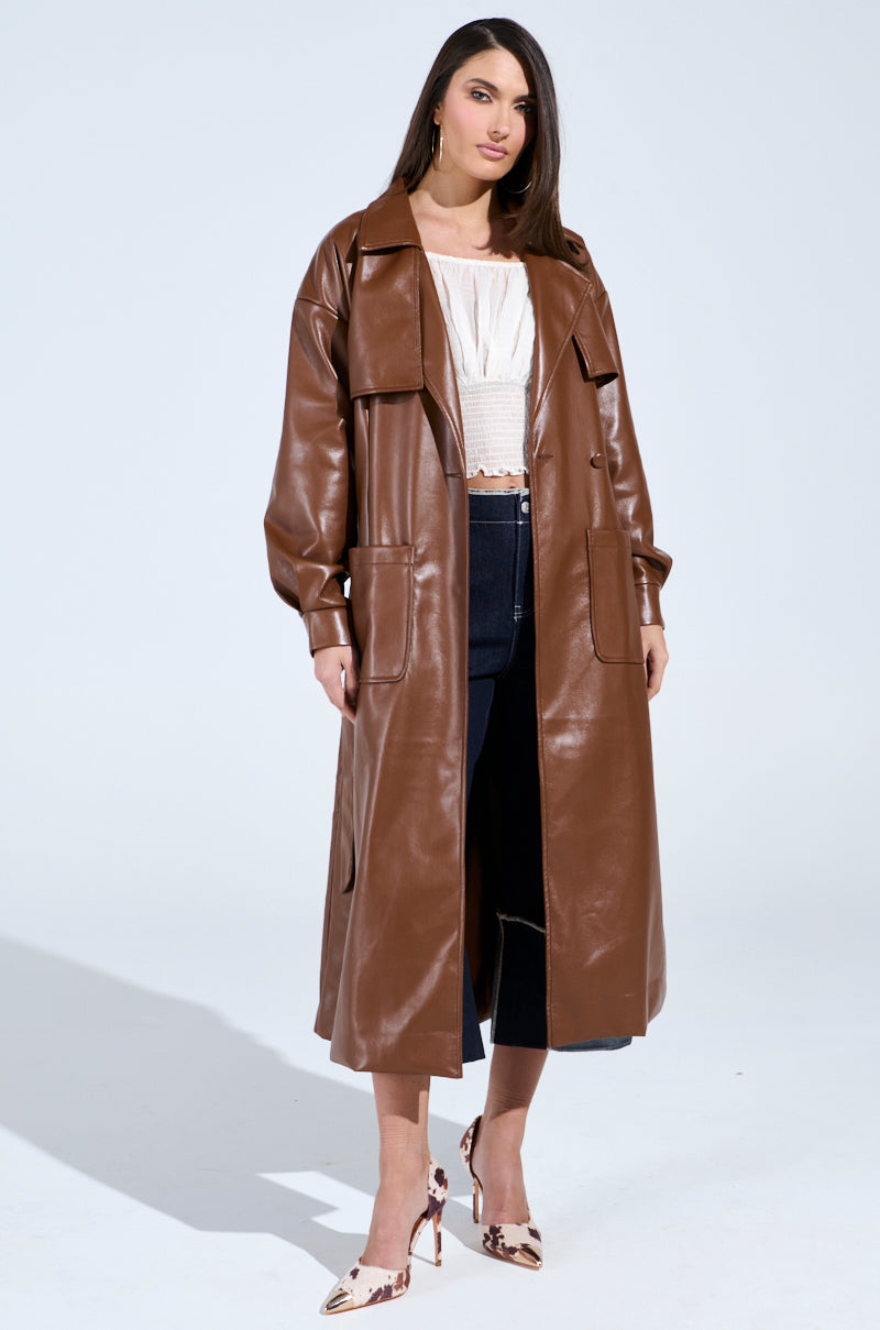 A woman exudes confidence against a plain backdrop, donning the BUTTER LUXE FAUX LEATHER TRENCH IN BROWN over a white top and dark jeans, finished with high heels. Her trench coat offers timeless elegance with a modern flair.