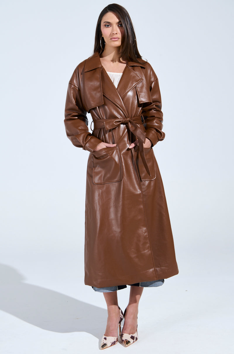 Against a plain background, a person models the BUTTER LUXE FAUX LEATHER TRENCH IN BROWN, cinched at the waist with its belt. Their shoulder-length hair adds flair to the chic look, completed by bold leopard print pointed-toe shoes.