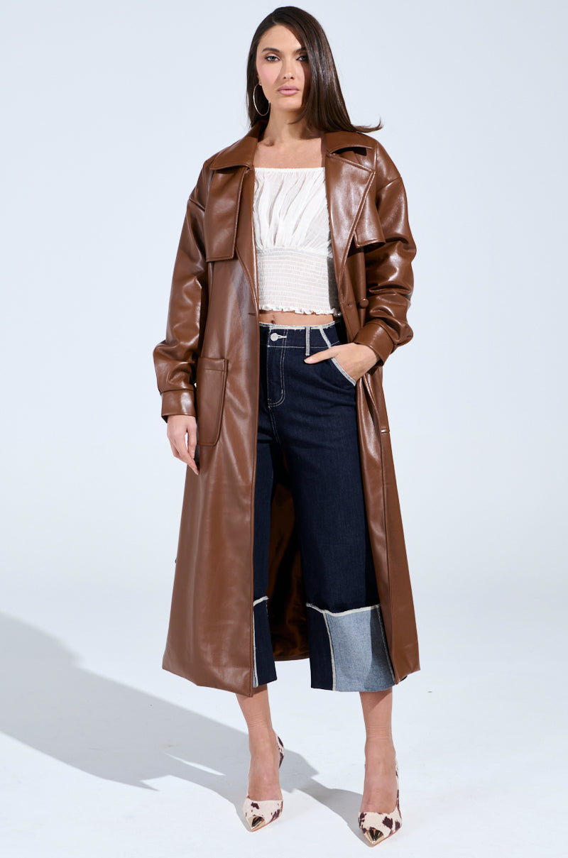 In a plain setting, an individual exudes confidence in the BUTTER LUXE FAUX LEATHER TRENCH IN BROWN over a white textured top and dark wide-leg jeans with light denim cuffs. They accessorize with hoop earrings and patterned high heels, with one hand casually tucked into their pocket.