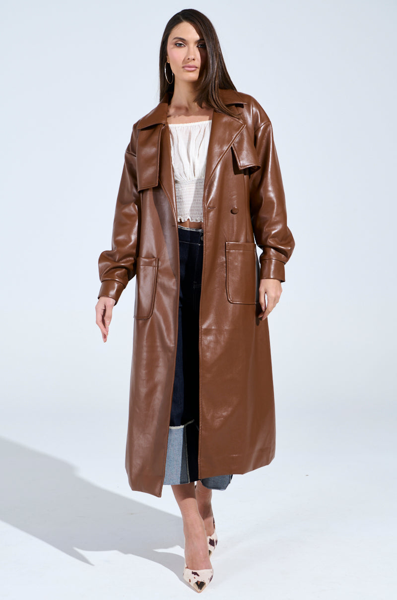 A woman poses confidently in a sophisticated BUTTER LUXE FAUX LEATHER TRENCH IN BROWN, complemented by a white blouse and dark pants. She wears hoop earrings, pointy white shoes, and her straight hair cascading over one shoulder against a plain light background.