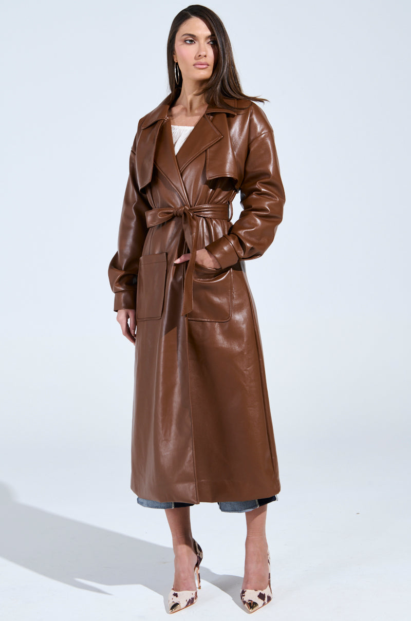 A woman confidently stands in a BUTTER LUXE FAUX LEATHER TRENCH IN BROWN over a white top and cropped jeans, complemented by animal print high heels against a plain, light background.