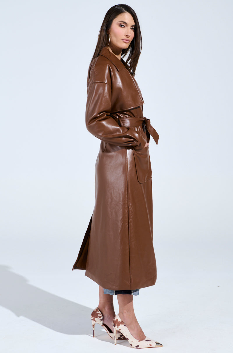 A woman exudes confidence in a Butter Luxe faux leather trench in brown with a belt, perfectly paired with light blue jeans. Her long dark hair flows as she poses sideways, showcasing her beige pointed shoes against a plain white background.