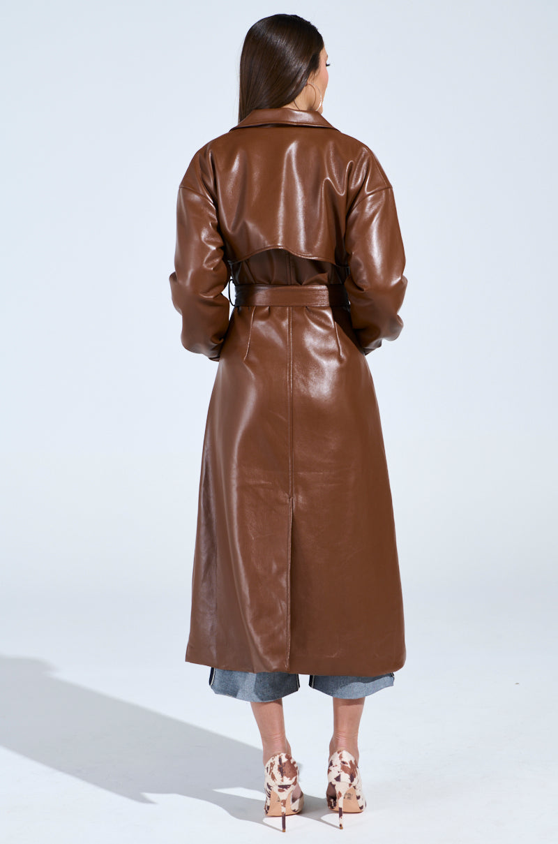 A person with long hair, facing away, wears the BUTTER LUXE FAUX LEATHER TRENCH IN BROWN belted over blue jeans. Their high heels feature a cow-print design, set against a plain light gray background highlighting this stylish ensemble.