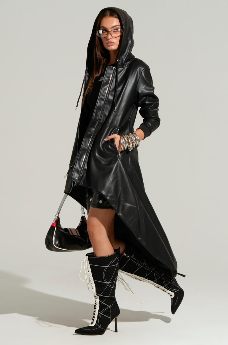 A person wearing the NEW YORK MINUTE HIGH LOW TRENCH, a long black faux leather jacket with a hood, paired with a black shirt, black skirt, and stylish black and white lace-up knee-high boots. They also wear glasses and hold a black handbag, accessorized with several bracelets and rings.
