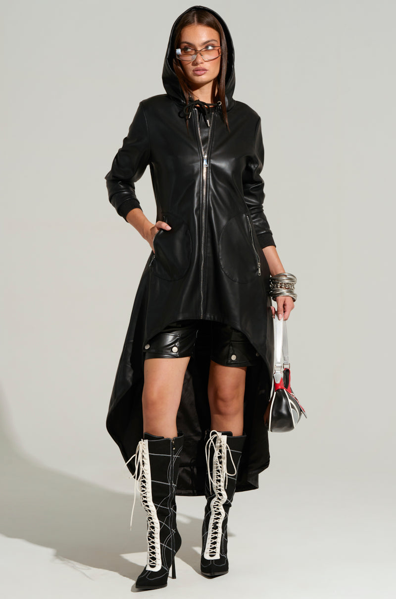A person wearing the NEW YORK MINUTE HIGH LOW TRENCH, a sleek black faux leather hooded jacket with a high-low hem, stands confidently. They pair it with black shorts and black knee-high boots featuring white laces. The person accessorizes with sunglasses, multiple bracelets, and a small handbag.