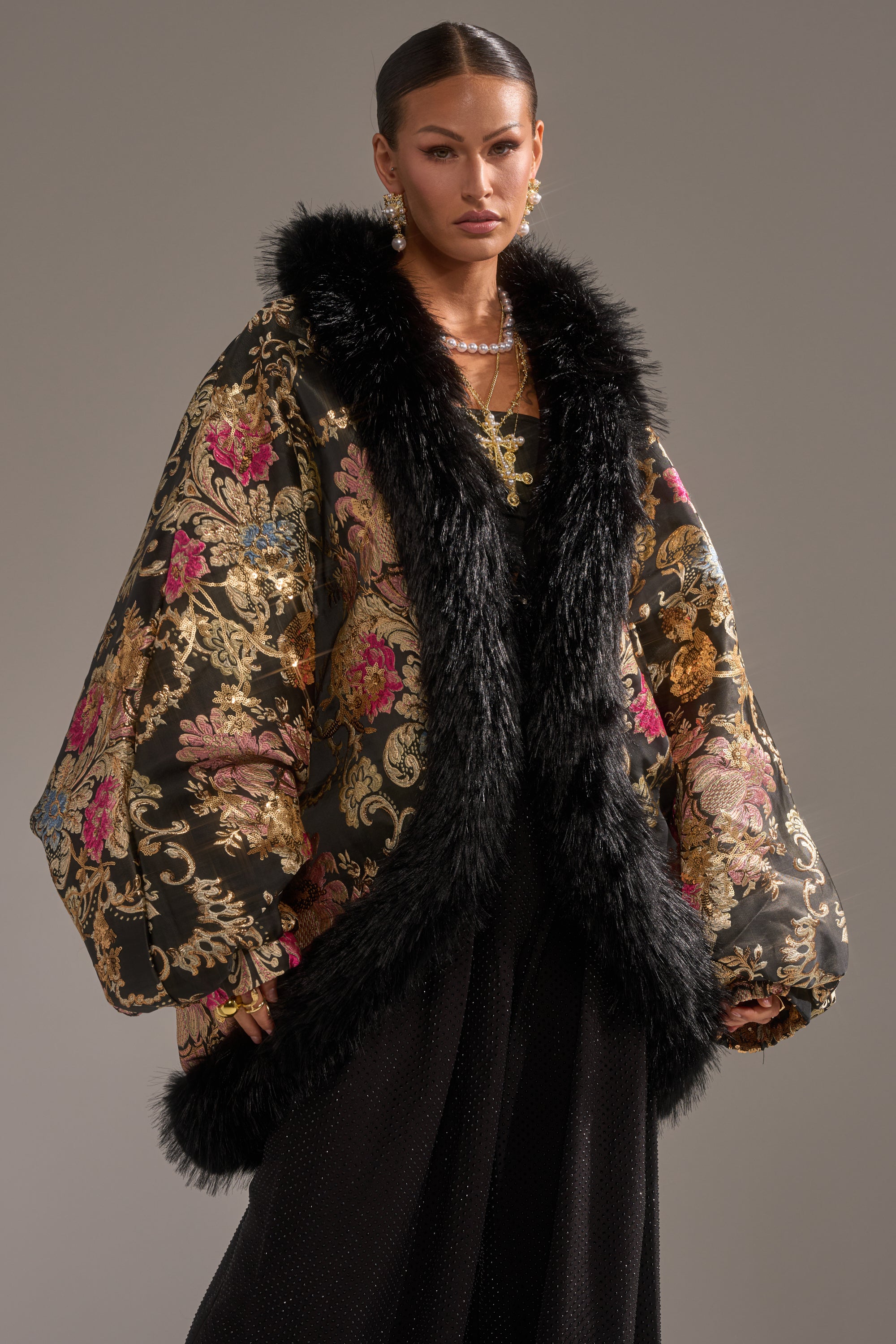 A woman models the LOLA COCOON COAT IN BROCADE, featuring an ornate gold, pink, and blue floral pattern with thick black faux fur trim. She pairs it with sleek hair, statement earrings, and layered necklaces against a plain background.