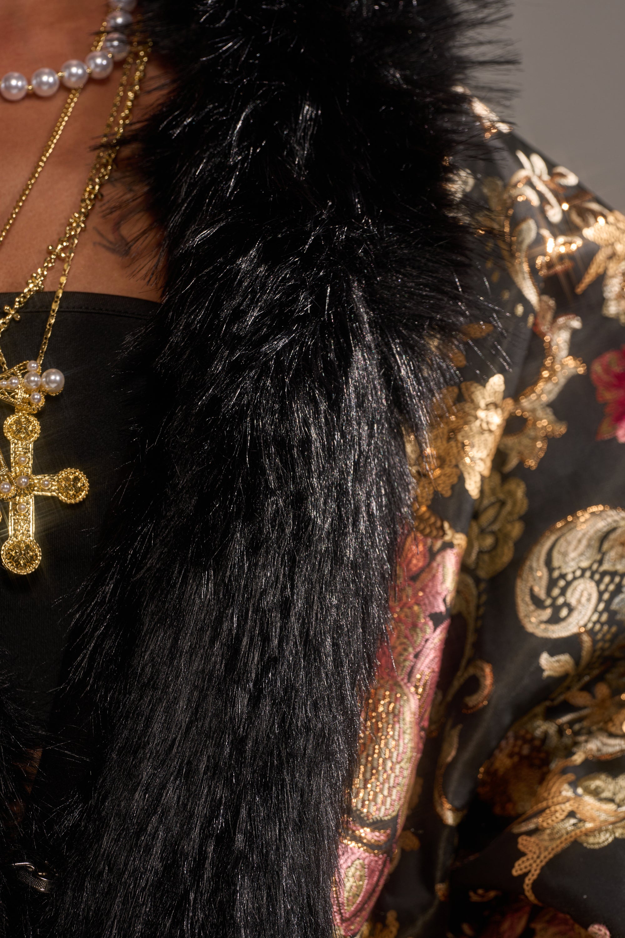 A close-up of a person wearing the LOLA COCOON COAT IN BROCADE, styled with a black fur shawl and gold and pearl necklaces featuring a cross pendant, over an ornate floral garment in gold, red, and green.