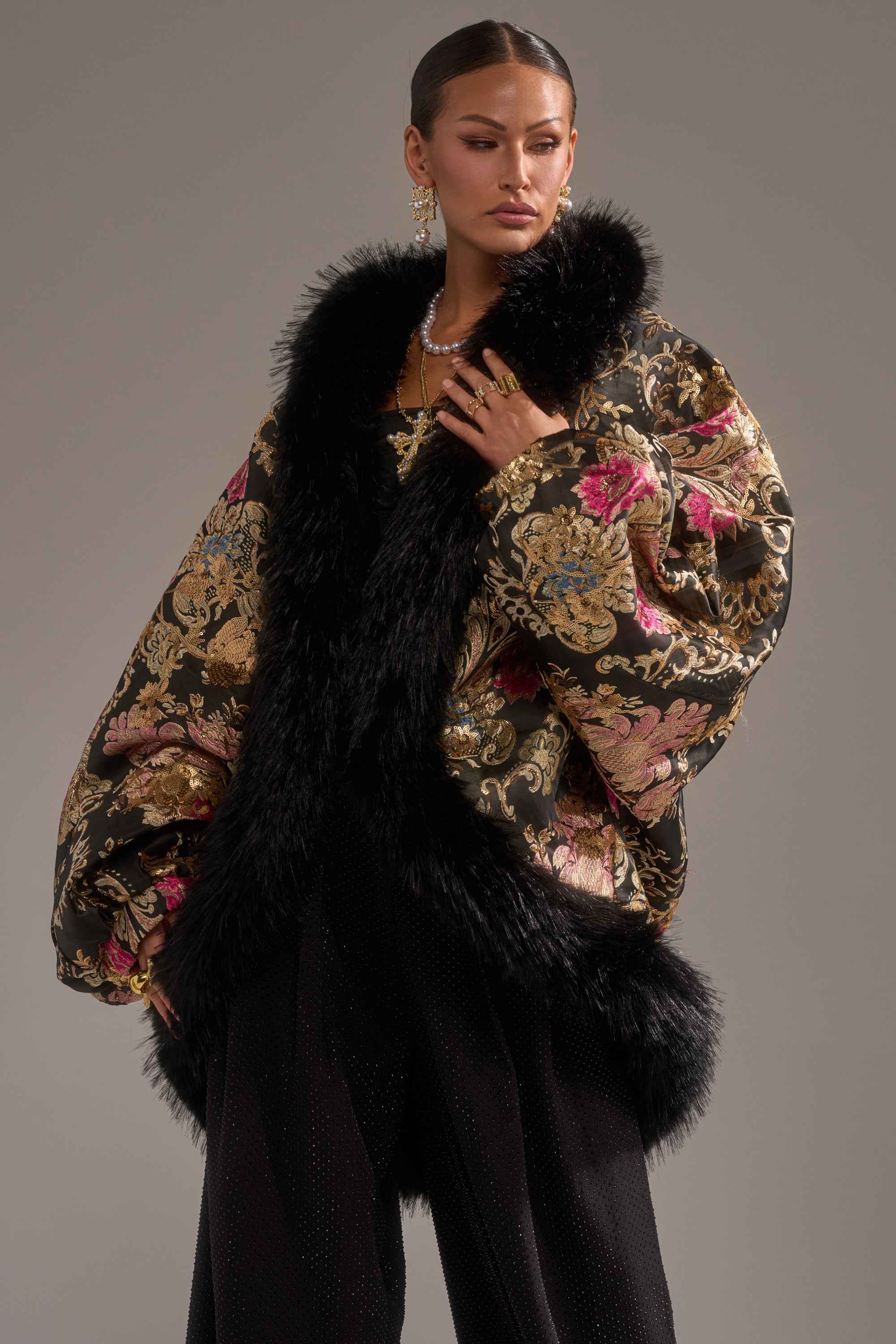 A woman models the LOLA COCOON COAT IN BROCADE, featuring luxurious black and gold fabric with a large black faux fur collar, paired with sparkling earrings and dark pants against a plain gray backdrop.