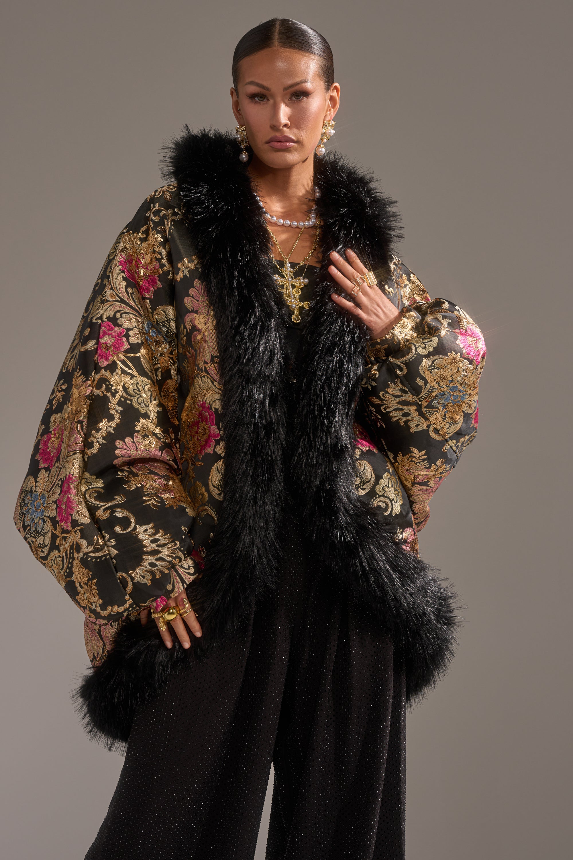 A woman poses confidently in the LOLA COCOON COAT IN BROCADE, featuring a floral pattern and black faux fur trim, styled with black wide-leg pants and gold jewelry against a plain background.