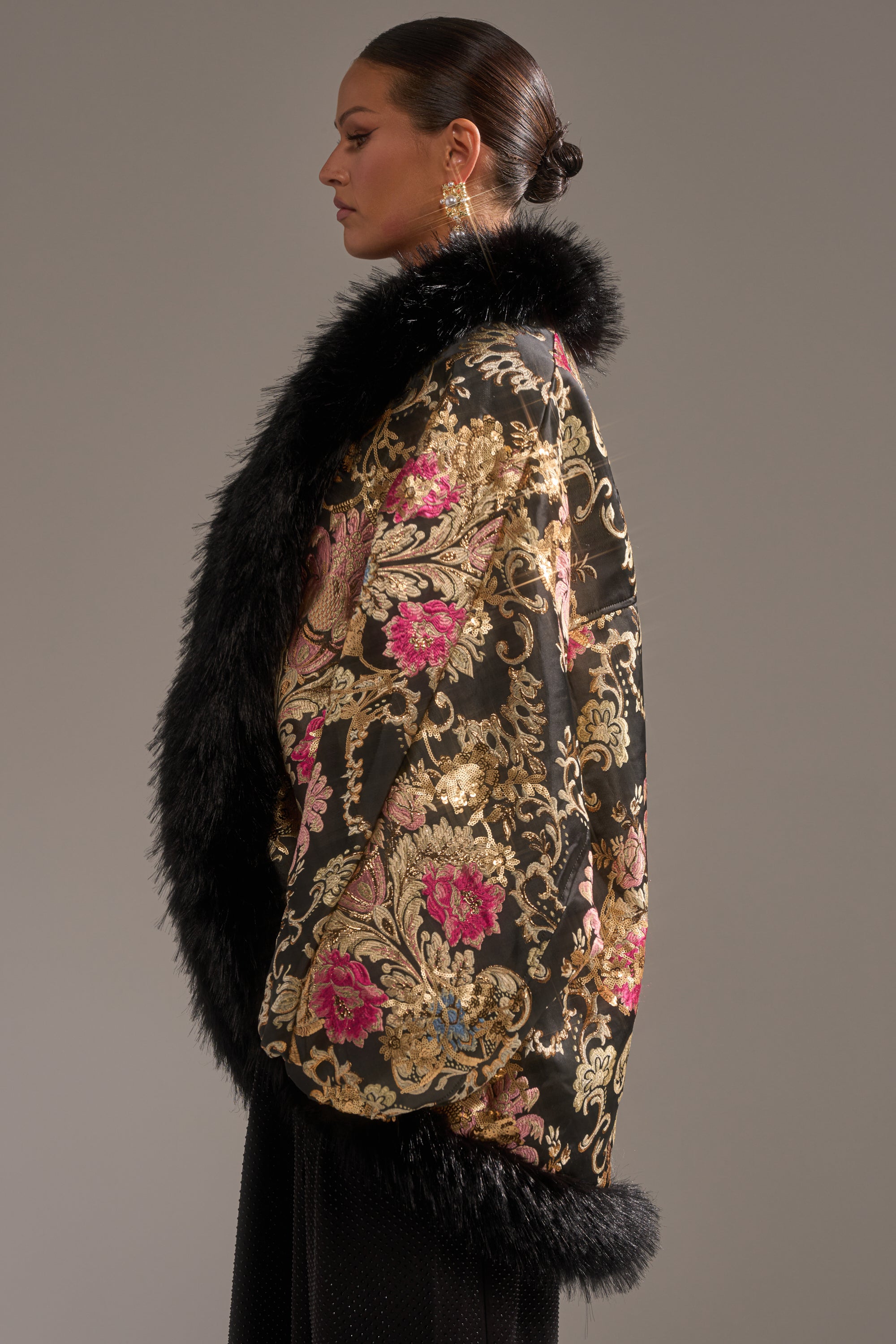 A woman in a sleek bun and ornate earrings poses sideways against a plain background, modeling the LOLA COCOON COAT IN BROCADE—a luxurious black cocoon coat with gold and pink floral embroidery and faux fur trim.