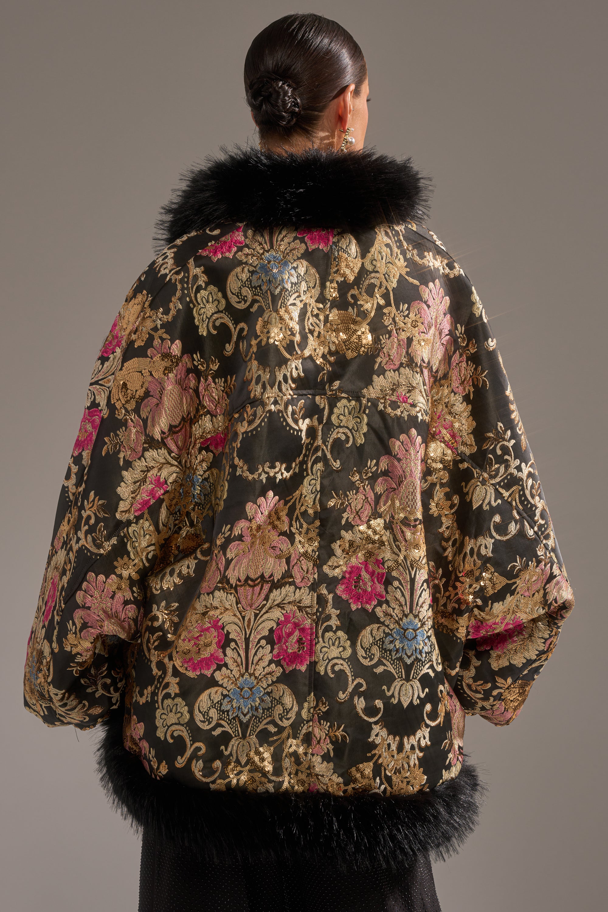 Seen from behind, a person with dark hair in a low bun wears the LOLA COCOON COAT IN BROCADE—luxurious black and gold brocade with pink and blue accents, plus black faux fur trim on the collar, cuffs, and hem, against a neutral background.