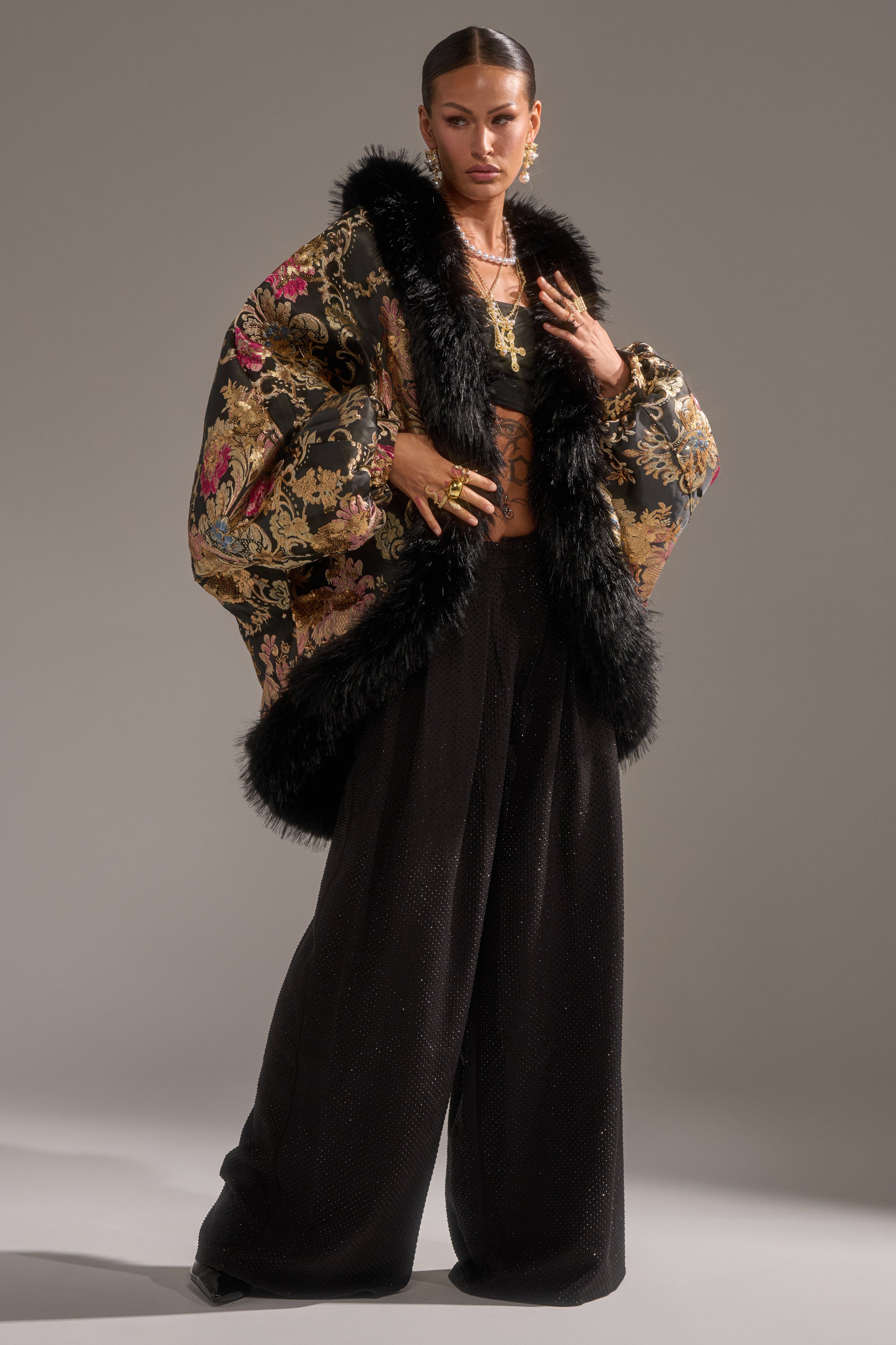 A person confidently poses against a gray background, wearing the LOLA COCOON COAT IN BROCADE with faux fur trim and wide black pants. Their slicked-back hair, bold jewelry, and neutral expression complete the look.
