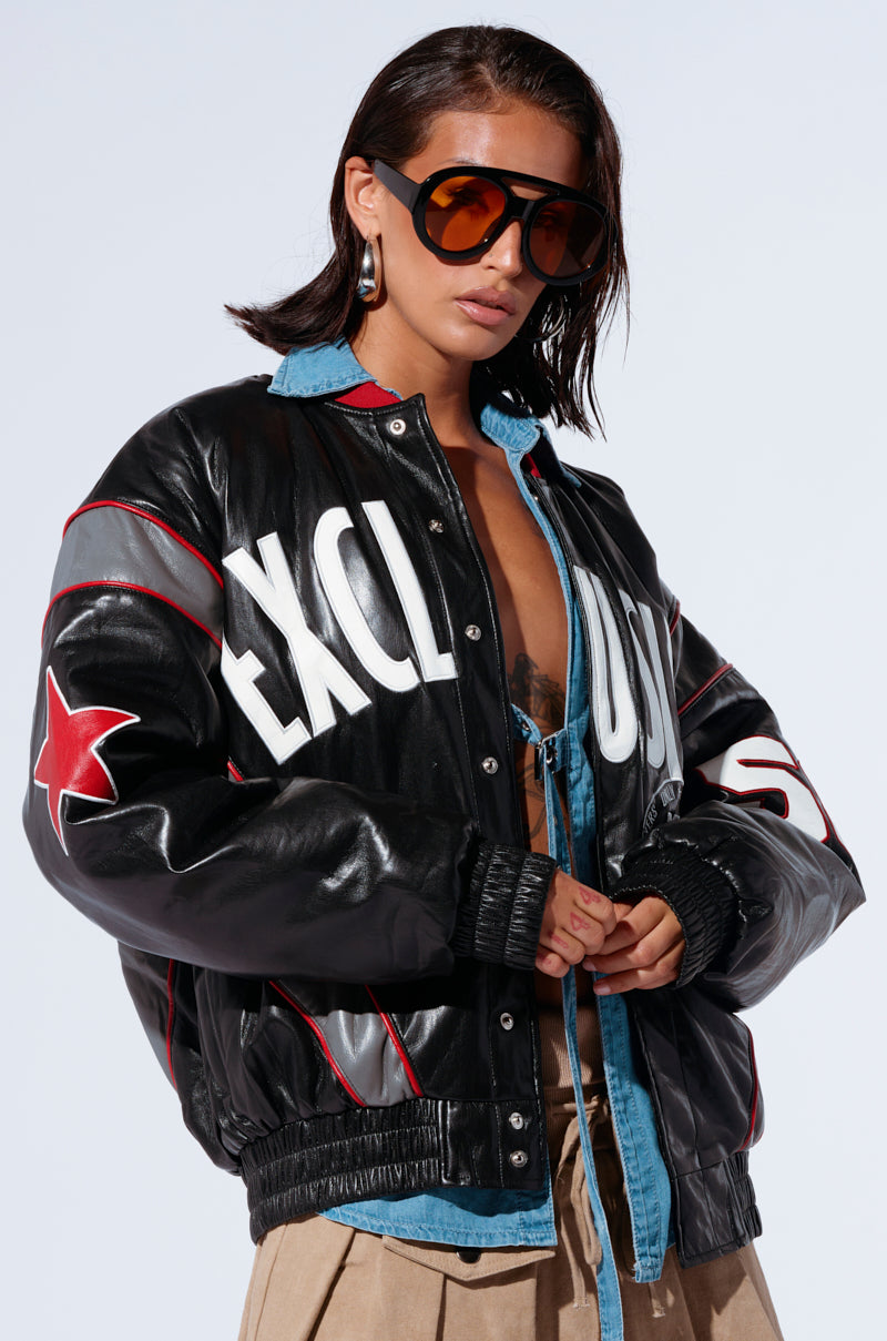 A person with shoulder-length dark hair wears large, amber-tinted sunglasses and a ROADRUNNER EMBROIDERED BOMBER with white letters and a red star emblem. The jacket is open, revealing a denim shirt underneath. The individual is also wearing beige pants.