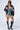 A person stands posing against a plain backdrop, wearing large sunglasses, the ROADRUNNER EMBROIDERED BOMBER varsity jacket in black faux leather with white text and numbers, a denim crop top, and a tan pleated mini skirt. They also have black knee-high lace-up boots.