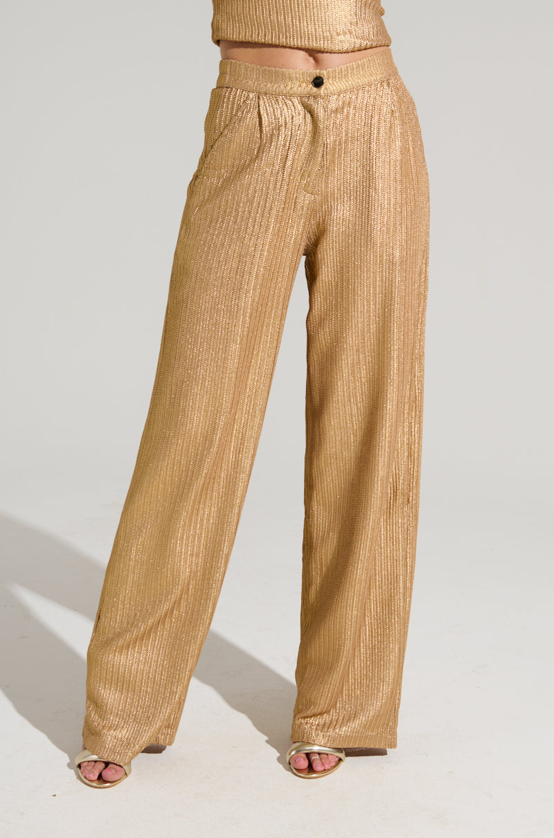 A person wearing the UP ALL NIGHT THINKING TROUSER IN GOLD and open-toed sandals stands against a plain background. The high-rise wide-leg trousers are loose and flowing, catching the light beautifully. The shot is cropped above the torso.