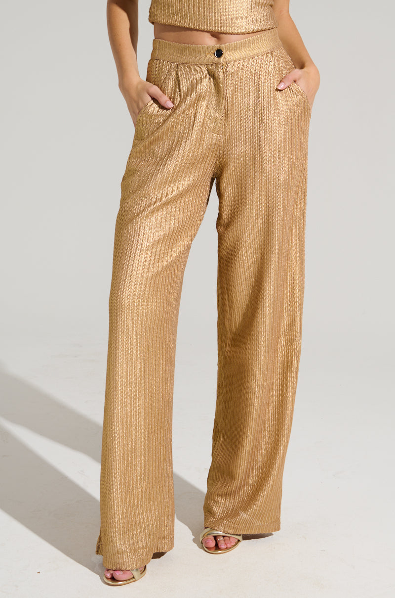A person is seen from the waist down, wearing the UP ALL NIGHT THINKING TROUSER IN GOLD, which features high-rise and wide-leg cuts with a slight shimmer. Their hands rest in the pants' pockets as they stand against a plain light-colored background, and the gold metallic trousers pair flawlessly with their open-toe sandals.