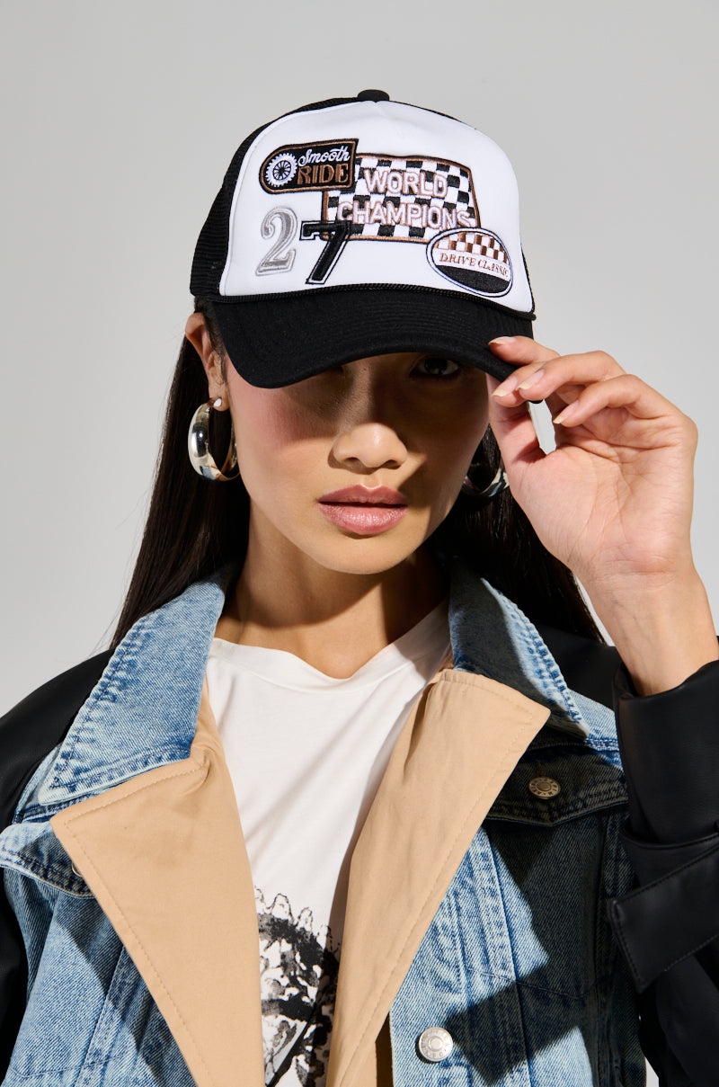 A person wearing a black and white WORLD CHAMPS PATCH TRUCKER HAT, featuring "World Champion 27" on the front. They have on a denim and leather jacket layered over a beige top, adjusting the hat with their right hand, while large hoop earrings add flair to their look.